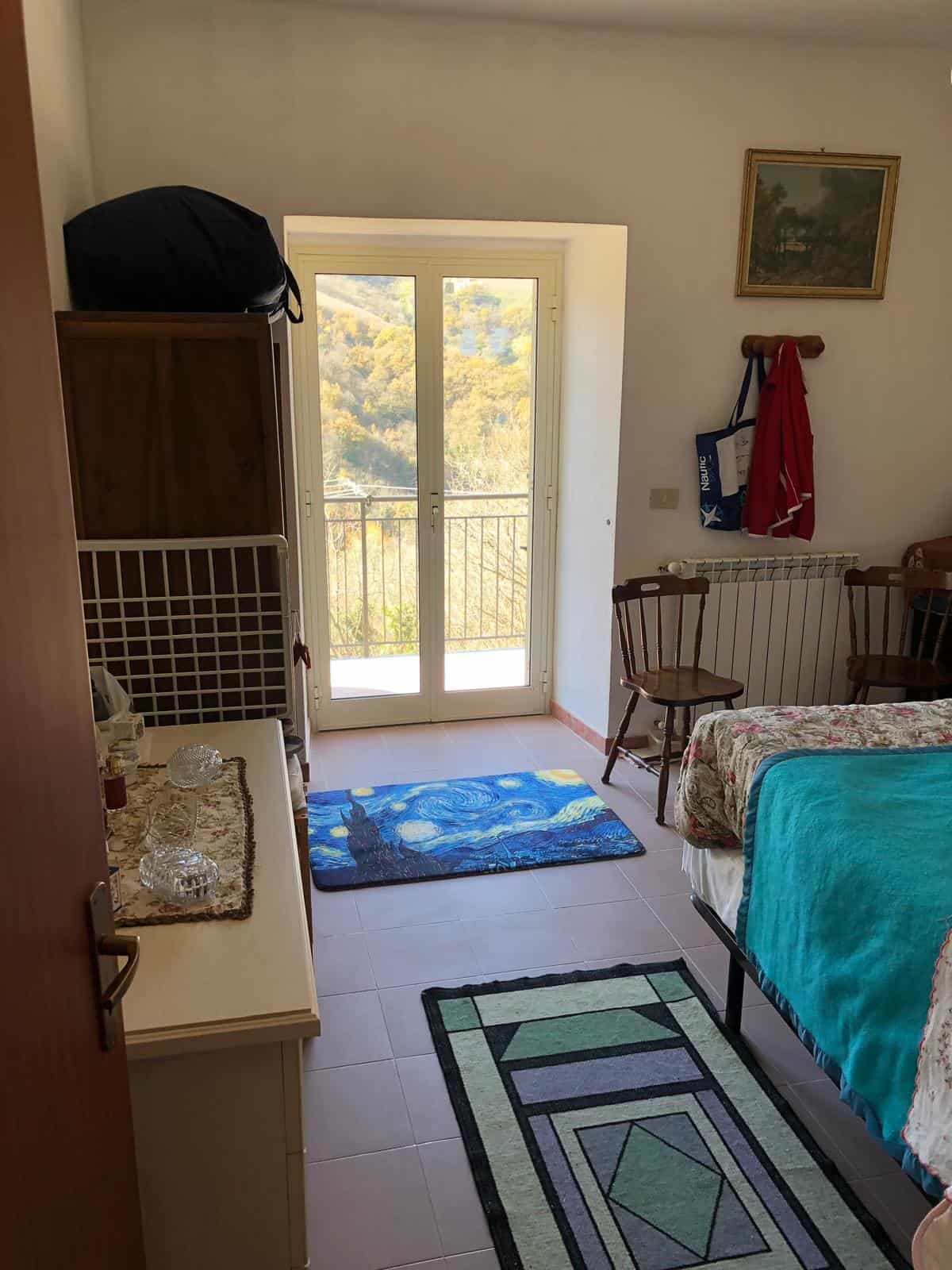Vista camera da letto con balcone panoramico, arredi classici, tappeto artistico, illuminazione naturale e mobili funzionali, ideale per vivere in tranquillità e godere della vista sulle colline e la natura circostante.