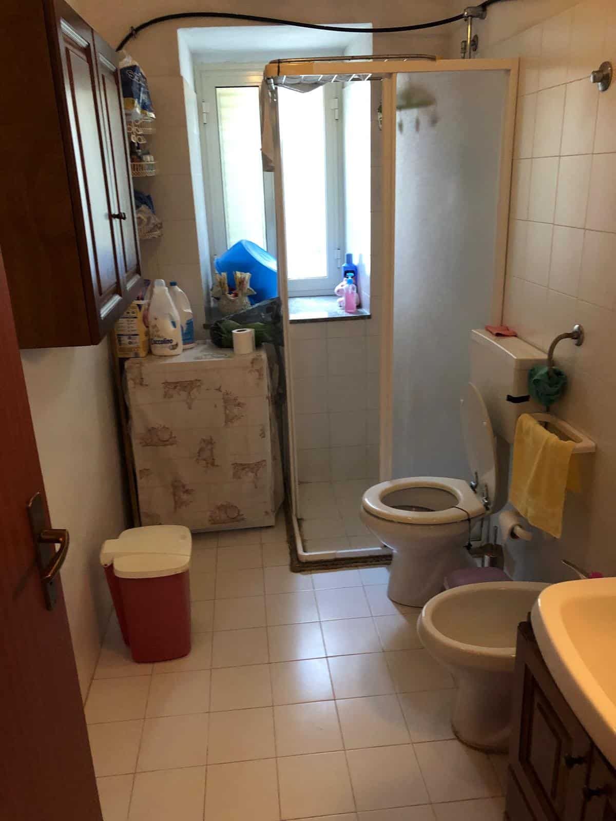 Spazio lavanderia bagno con wc e doccia, armadietto in legno, secchio rosso, vari prodotti per pulizia, finestra con luce naturale, ambiente funzionale e pratico.