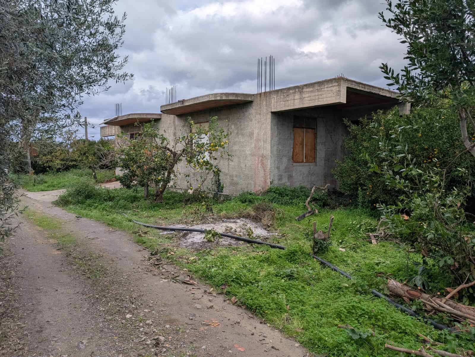 Rudere di casa in costruzione con struttura in cemento, circondata da alberi da frutto, su strada sterrata, cielo nuvoloso, ideale per progetto di ristrutturazione immobiliare.