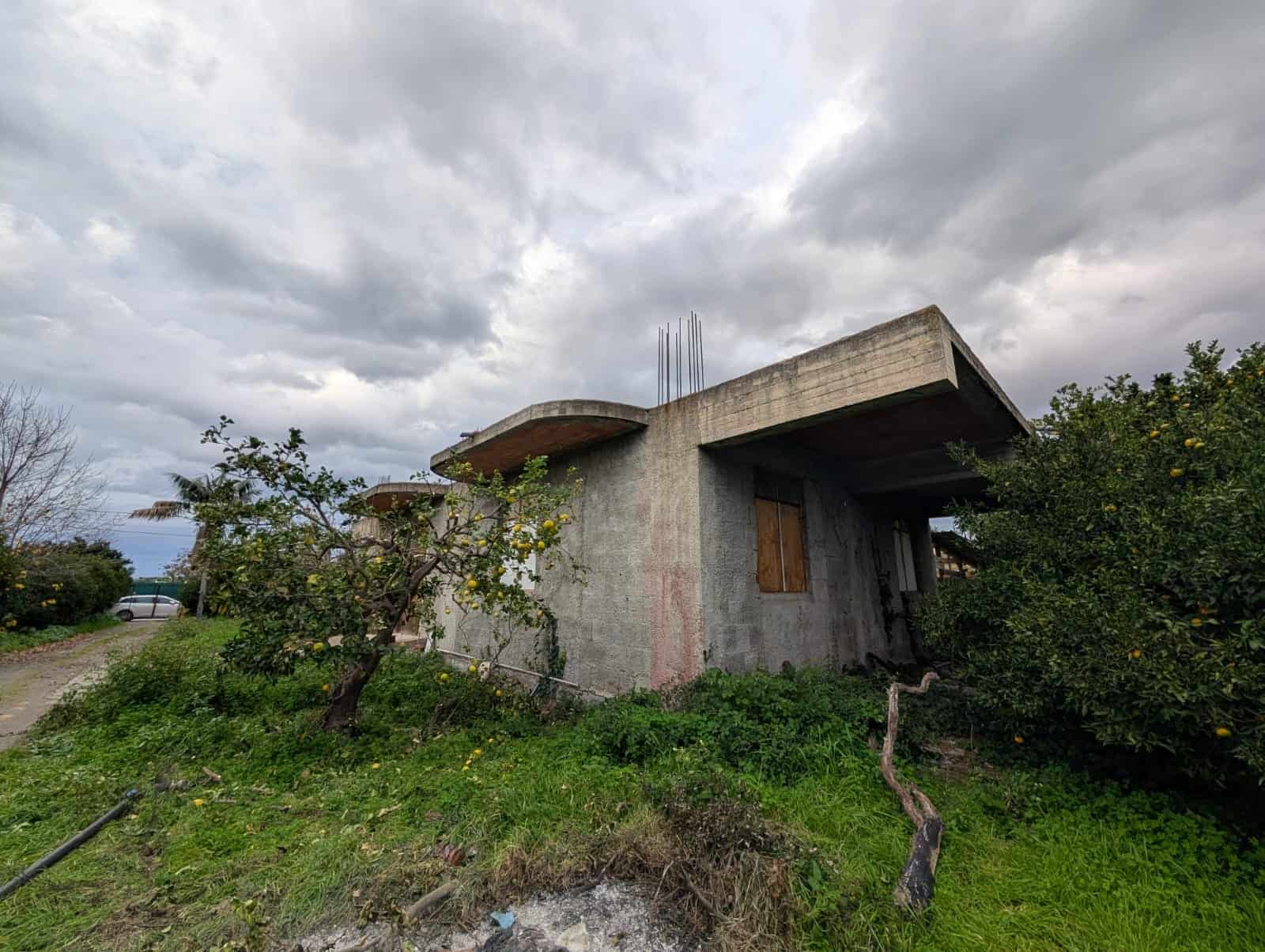 Casa in costruzione con struttura in cemento e giardino con alberi di limoni, sotto un cielo nuvoloso, pronta per essere trasformata in una casa moderna o tradizionale.