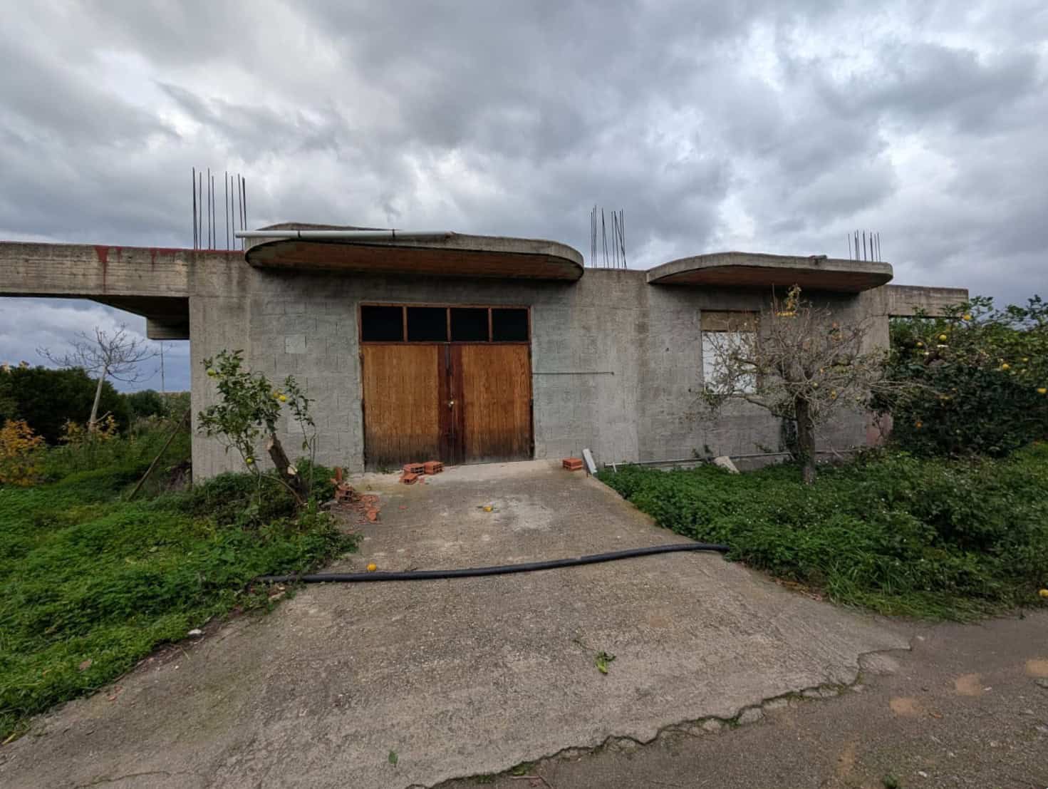Stabile in costruzione con pareti in cemento e infissi in legno, in sviluppo su un'area verde con alberi e piante, pronta per essere completata come casa privata o investimento immobiliare.