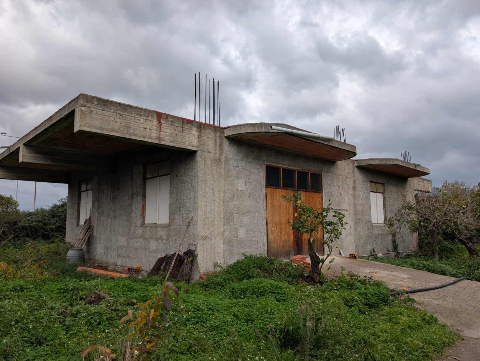 Casa in costruzione con struttura in cemento armato, finestre in vetro e portone in legno, situata in zona rurale con vegetazione e strada di accesso. Perfect real estate project, pronta a essere personalizzata.