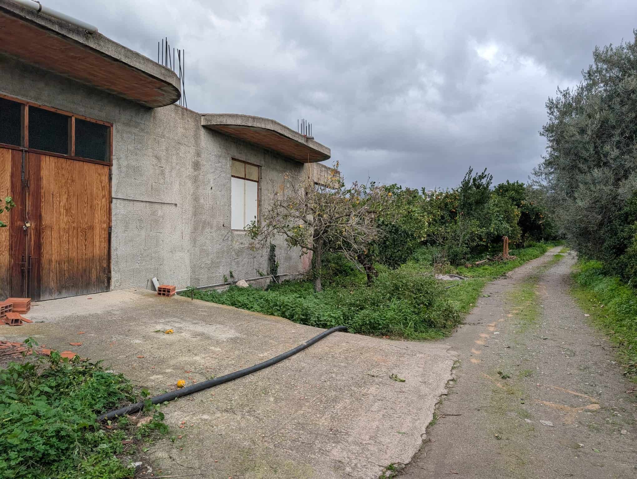 Vecchio edificio in costruzione con pareti in cemento e porte in legno, strada sterrata e natura circostante, progetto di ristrutturazione casa NextCasa, ambiente rurale e tranquillo, opportunità immobiliare.