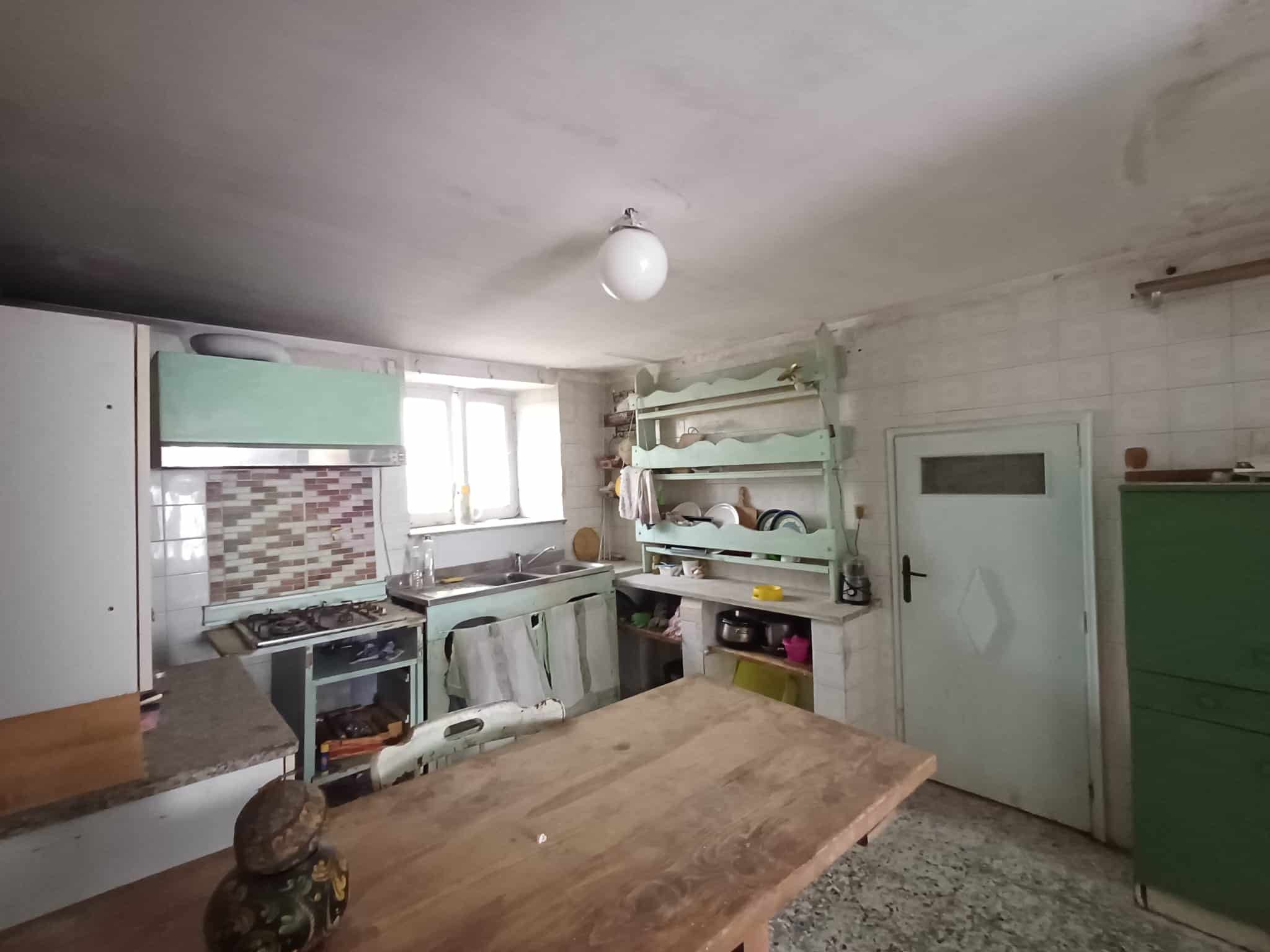 Il testo dell'immagine mostra una cucina rustica con mobili in legno, scaffali aperti e una finestra che lascia entrare luce naturale, in un ambiente semplice e funzionale.