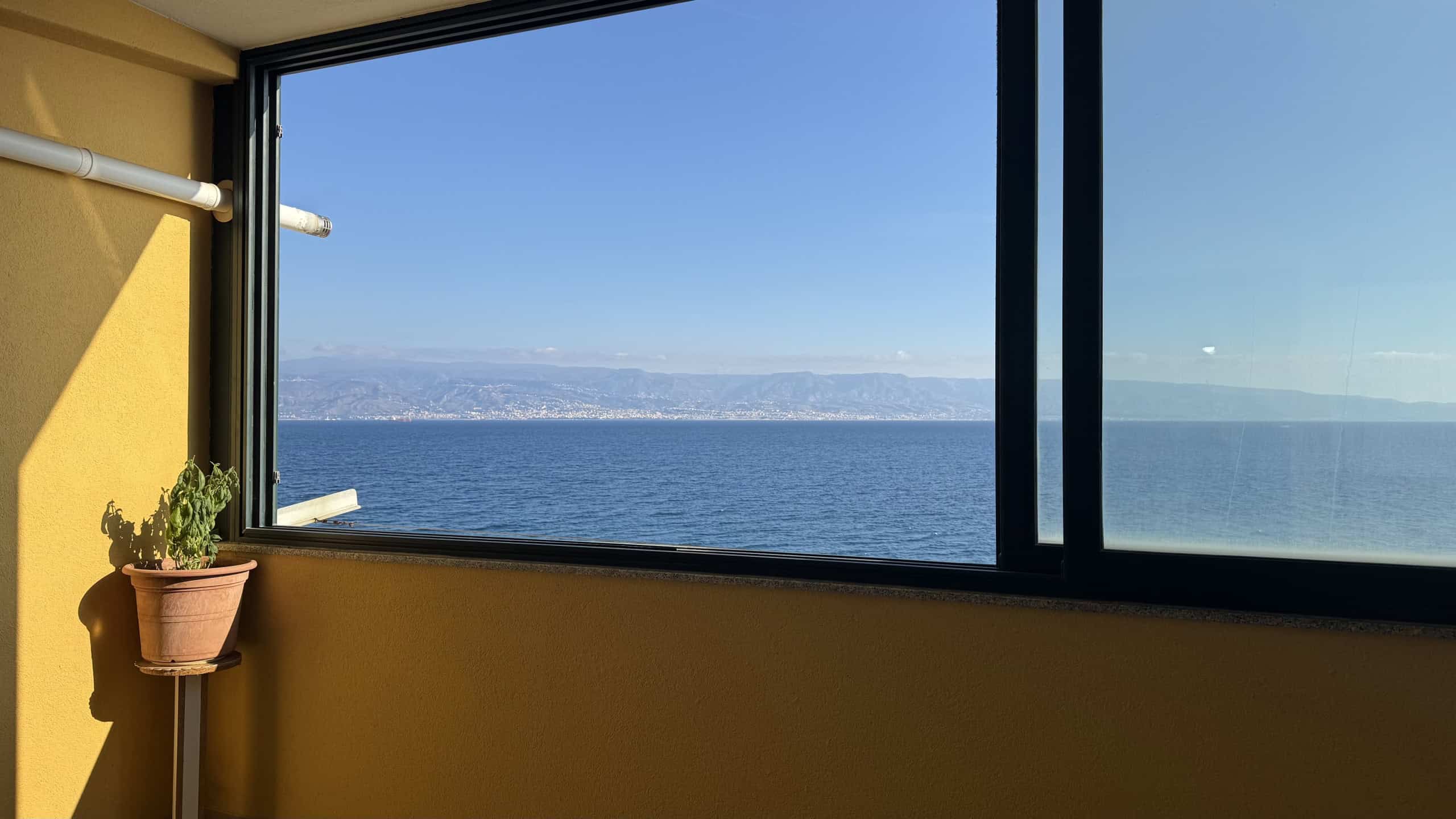 Vista mare dall'appartamento con ampia finestra, cielo sereno e montagne sullo sfondo, all'interno di una stanza con pareti gialle e pianta decorativa, ideale per immobili residenziali e investimento.