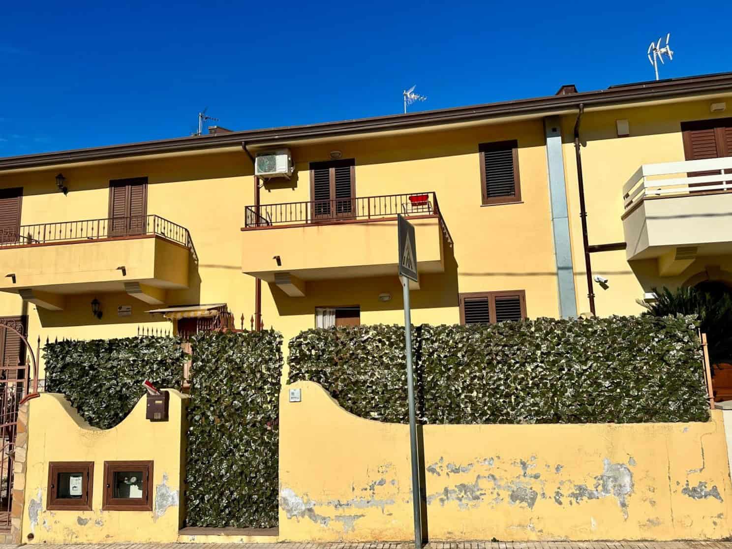 Casa in stile mediterraneo con balconi, finestre con persiane e recinzione verde, sotto un cielo blu, ambientazione residenziale italiana in contesto urbano.