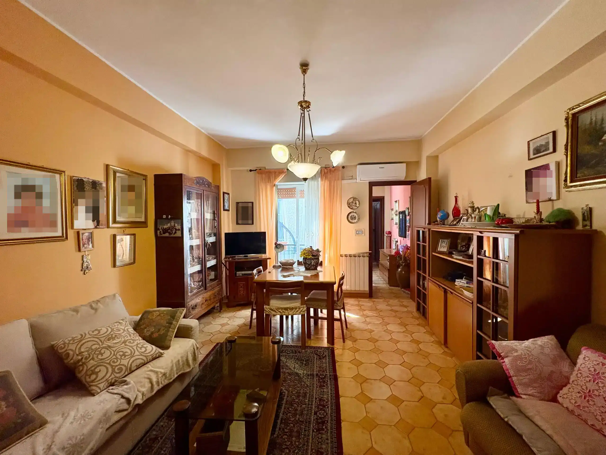 Ampio soggiorno con mobili in legno, pareti color panna, arredamento accogliente, lampadario centrale, cucina visibile in fondo, e accesso a balcone luminoso. Perfetto per vivere in comfort e stile.