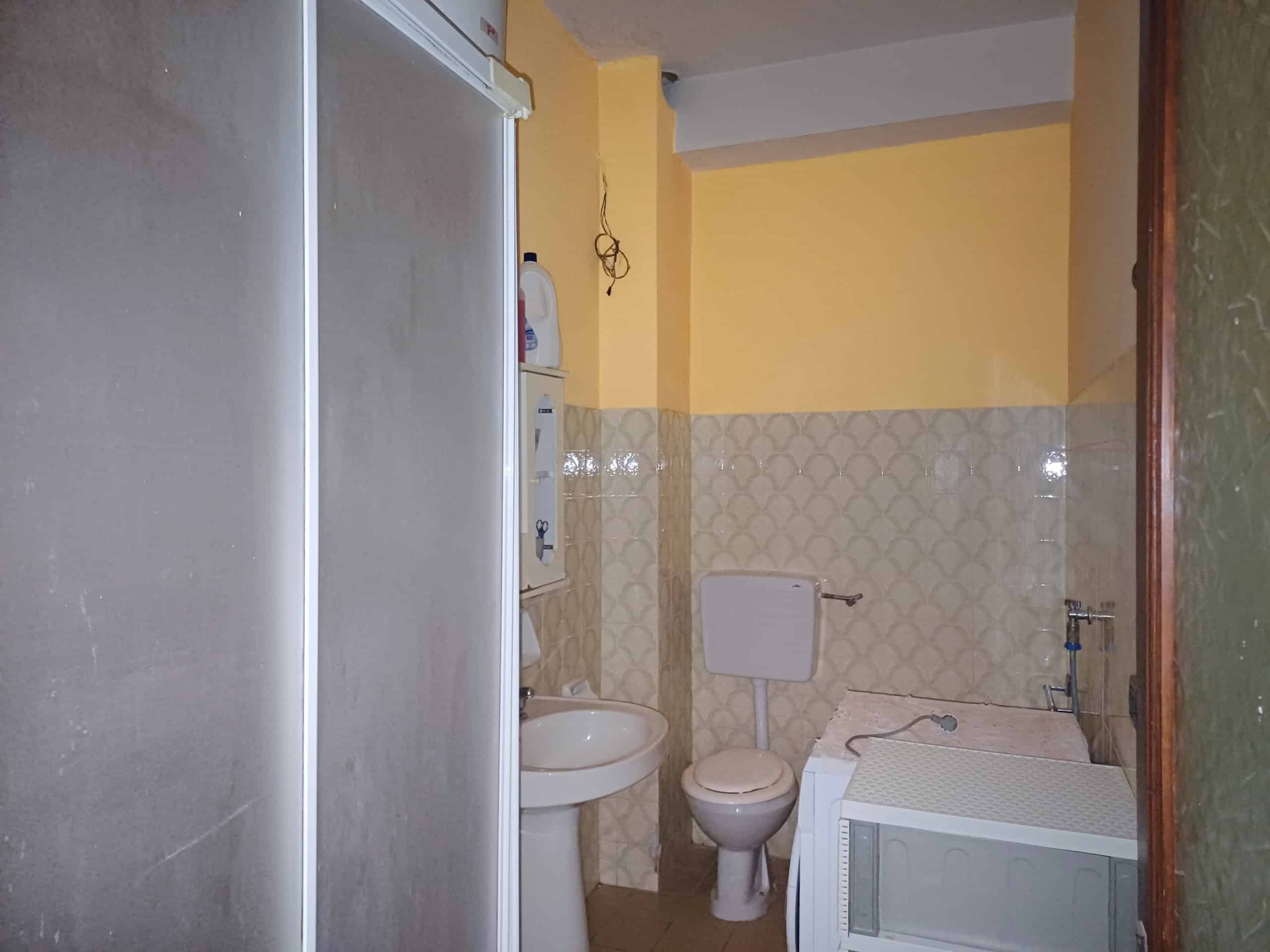 Bagno di piccole dimensioni con sanitari utilizzati, pareti rivestite con piastrelle beige e pareti gialle, componenti di arredamento essenziali, spazi sfruttati per funzioni di igiene quotidiana.