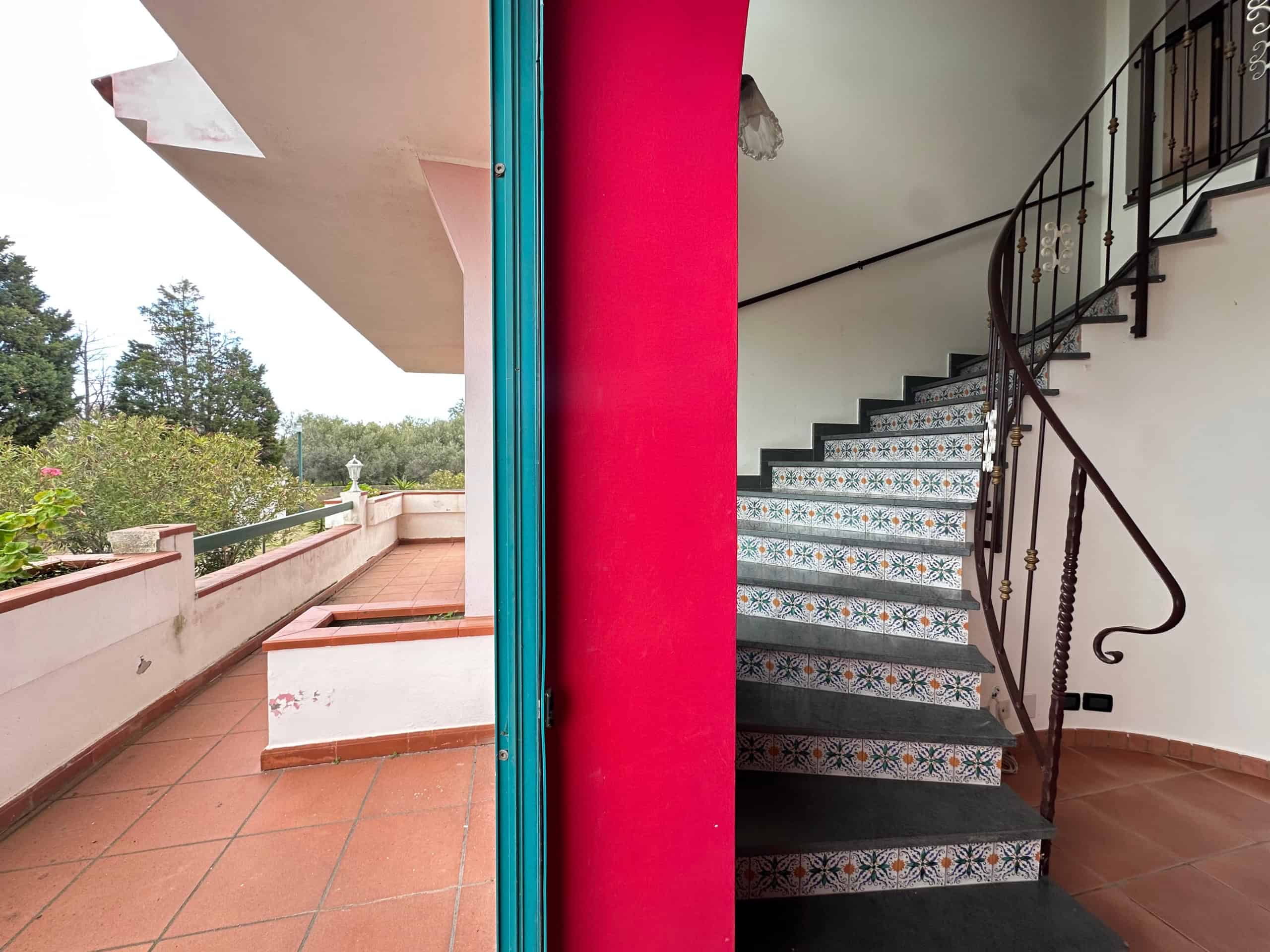 Spazio esterno e scala interna di una casa con dettagli colorati e decorativi, balcone con piante e vista sul verde. Perfetto esempio di ambienti resistenti, funzionali ed estetici per la casa.