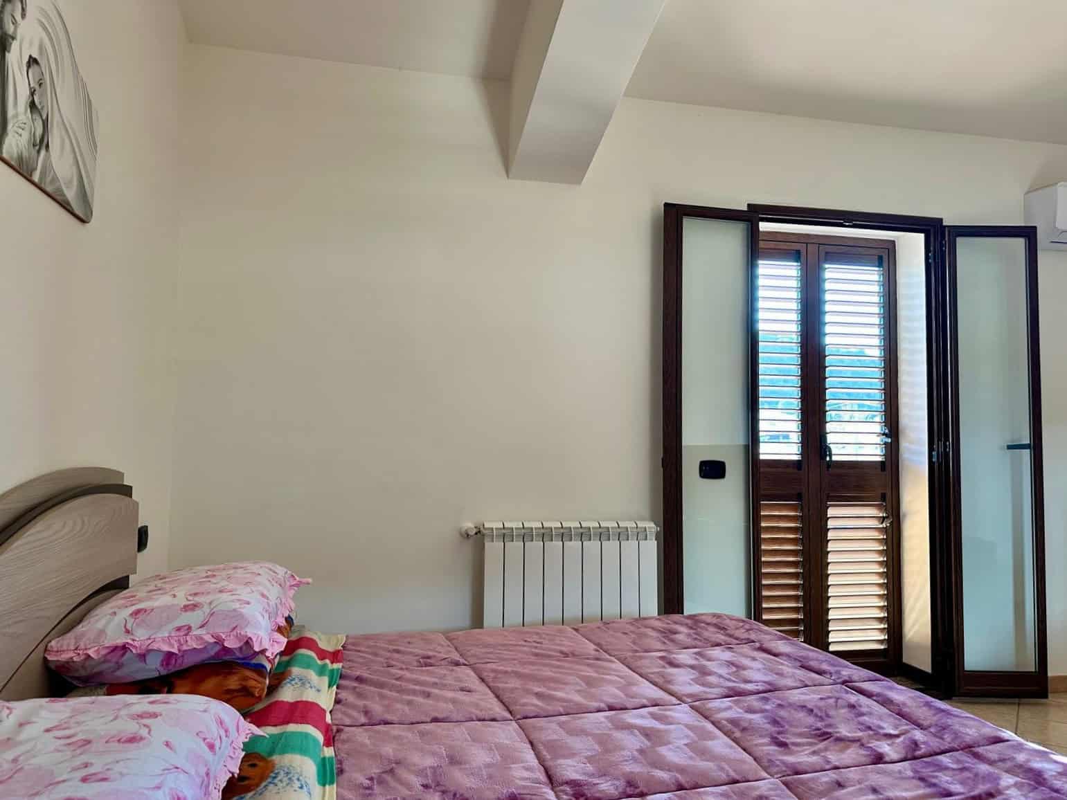 Accogliente camera da letto con letto matrimoniale, cuscini decorativi e comodini, finestra con persiane in legno e porta finestra che permette l'accesso al balcone, ambiente luminoso e confortevole.