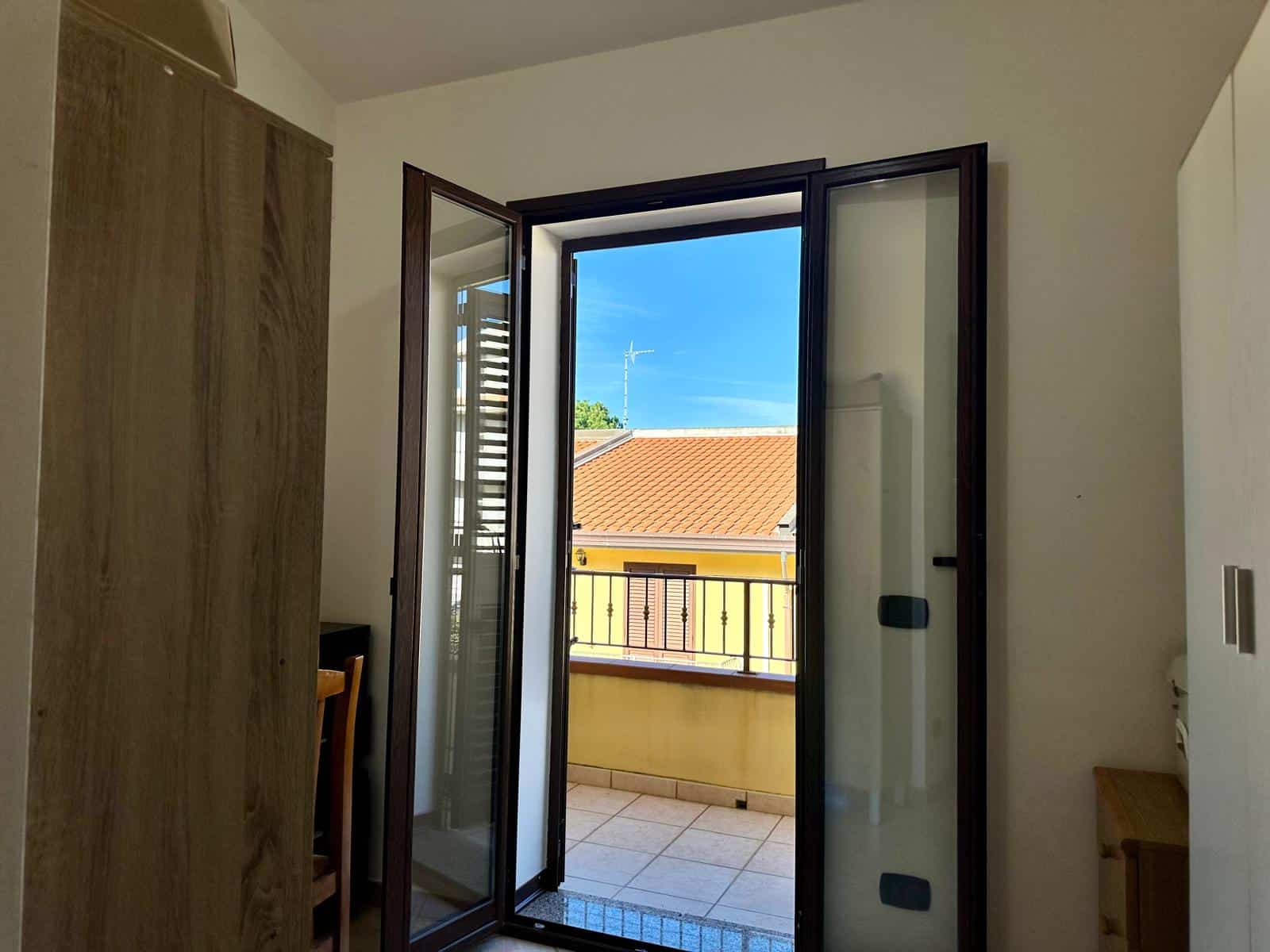 Balcone con porta finestra in legno e vetro, vista cielo blu e tetto in tegole rosse.