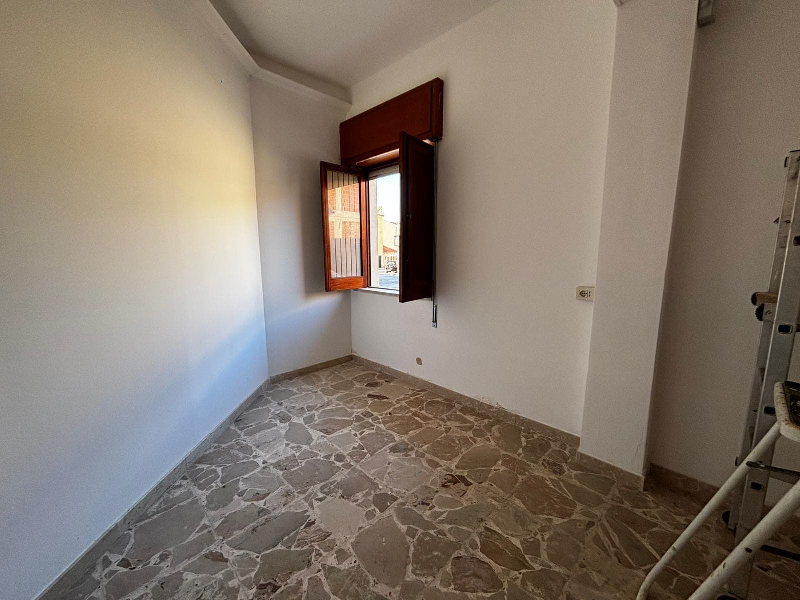 Stanza vuota con pareti bianche e finestra in legno, pavimento in marmo chiaro, illuminata dalla luce naturale, pronta per essere arredata e personalizzata.