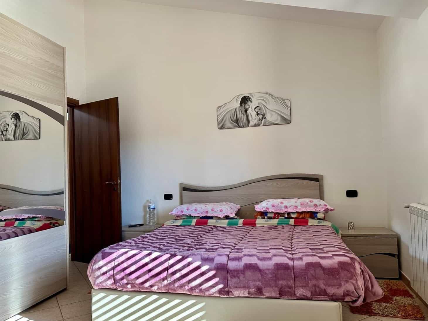Comodino in camera da letto matrimoniale con decorazione moderna, lenzuola rosa, mobili in essenza chiara e pareti bianche, ambientazione luminosa e accogliente.