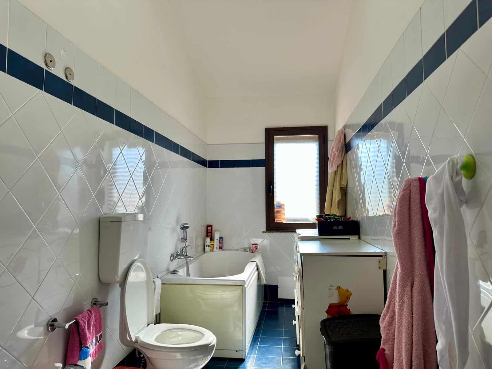 Lavabo e vasca da bagno in bagno luminoso con finestra, pareti rivestite in piastrelle bianche e blu, oggetti di uso quotidiano, asciugamani colorati e dettagli pratici per la cura personale.
