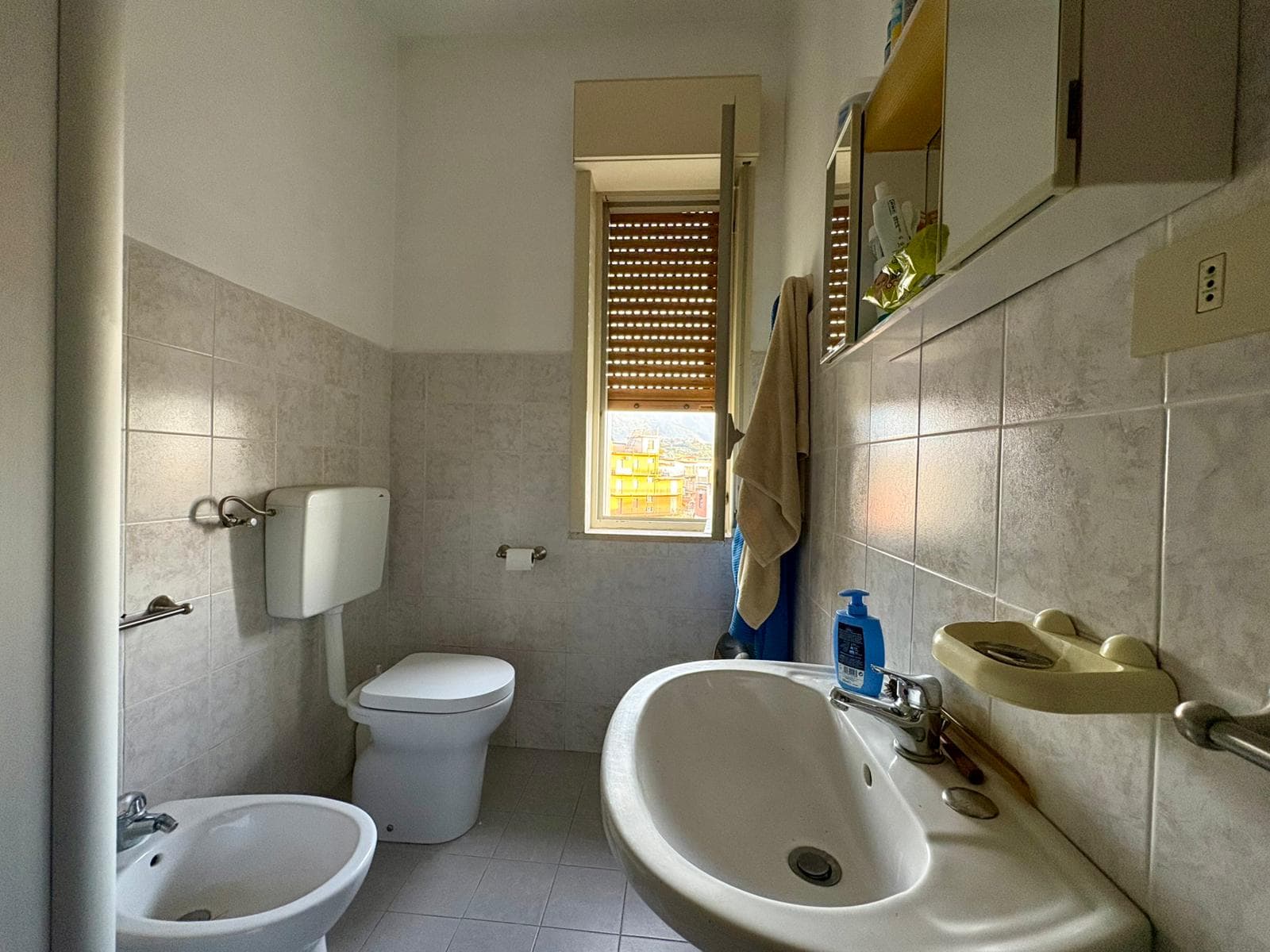 Lavabo, bidet e wc in un bagno con finestra e luce naturale, piastrelle chiaro, stile semplice e funzionale, ideale per appartamenti in vendita o affitto.