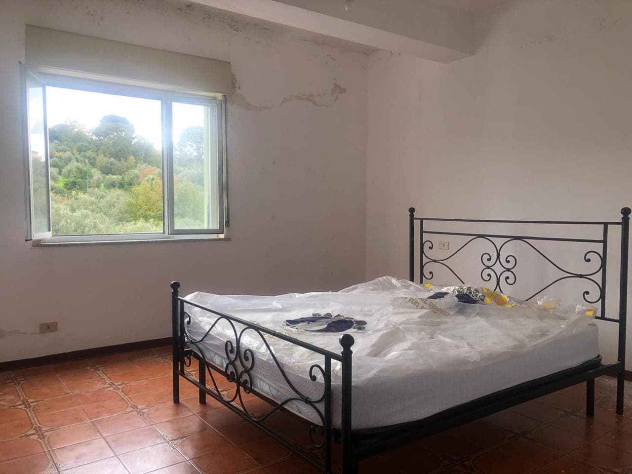 Arredamento camera da letto con letto in ferro battuto, finestra panoramica che affaccia su paesaggio verde, spazio semplice e luminoso, ideale per ristrutturazione o miglioramento abitativo.