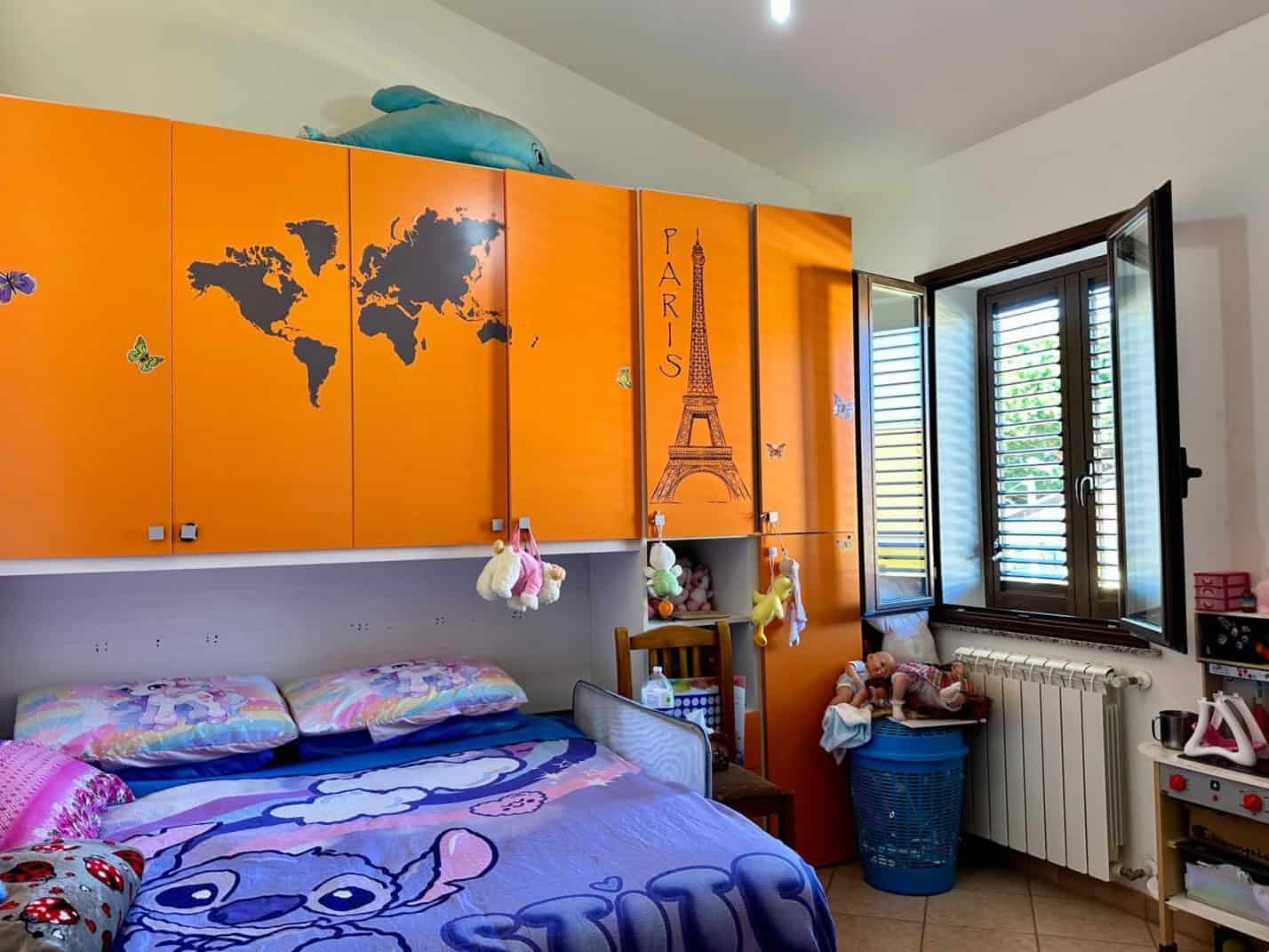 1. Cameretta bambini con decorazioni a tema Parigi, armadio arancione con mappa e Torre Eiffel, finestra con persiane, ambiente vivace e accogliente.