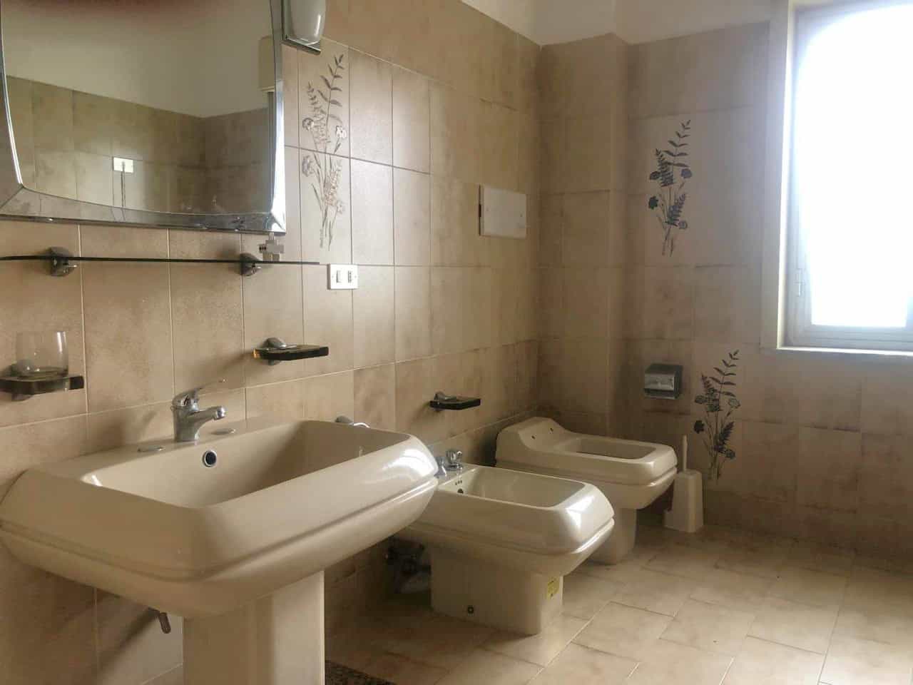 Lavabo e sanitari eleganti in bagno spazioso con pareti rivestite di piastrelle beige con motivi floreali. Ampia finestra per luce naturale, ideale per proprietà immobiliari di qualità.