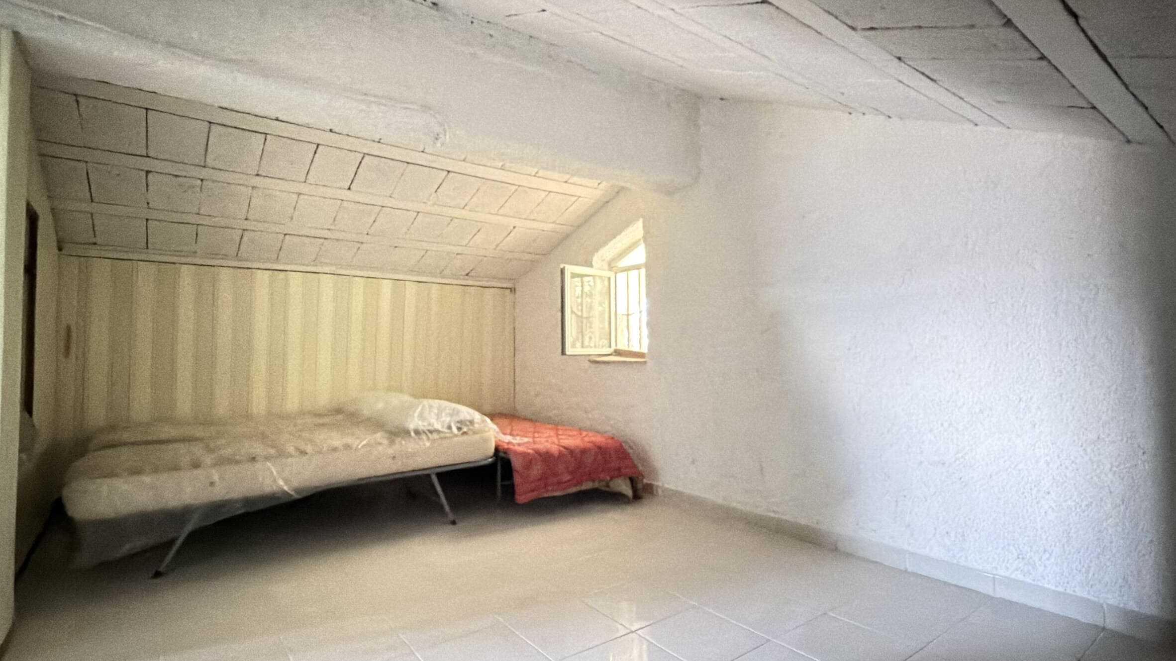 Sola stanza mansardata con letto e finestra, ideale per una camera da letto in un appartamento con soffitto inclinato e pareti in pietra bianca, ambiente luminoso e accogliente.