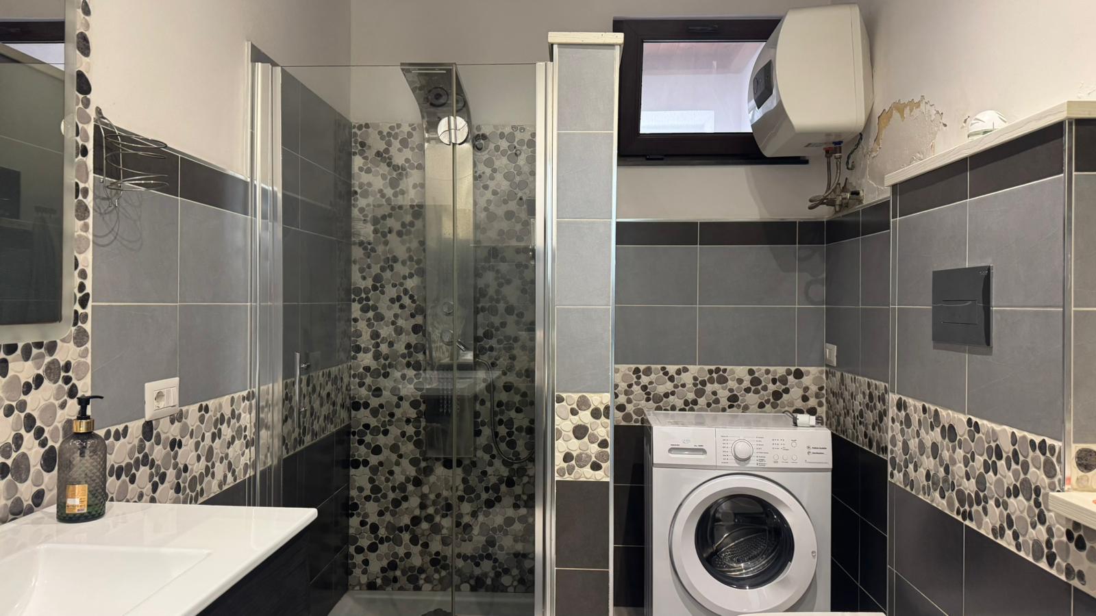 Lavanderia moderna con box doccia e lavabo elegante, pareti rivestite di piastrelle grigie e dettagli di mosaico colorato, con lavatrice e finestra per arieggiare.