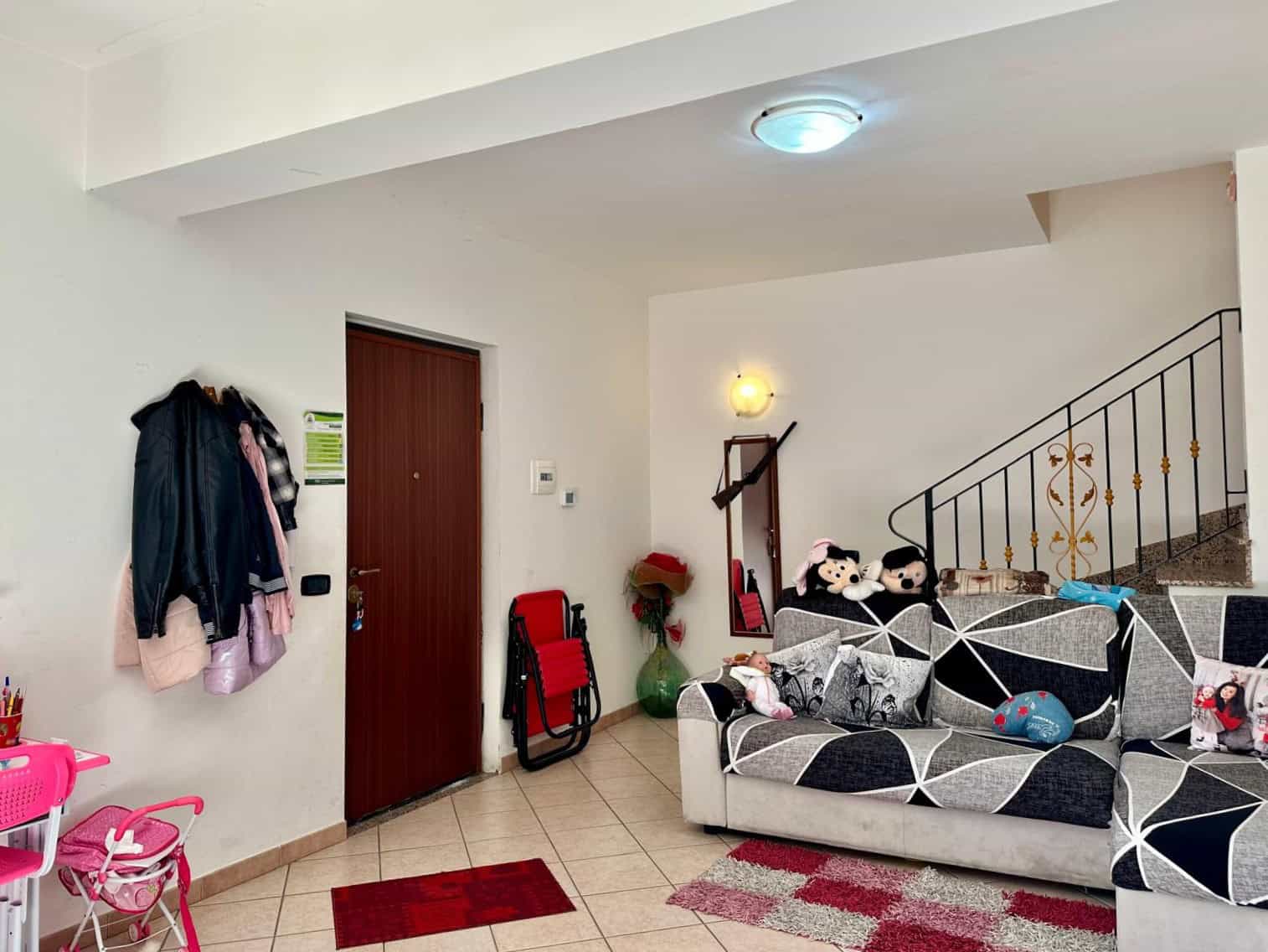Ingresso di appartamento arredato con divano, decorazioni per bambini, scale in ferro battuto, mobili funzionali, e ambiente accogliente in stile moderno.