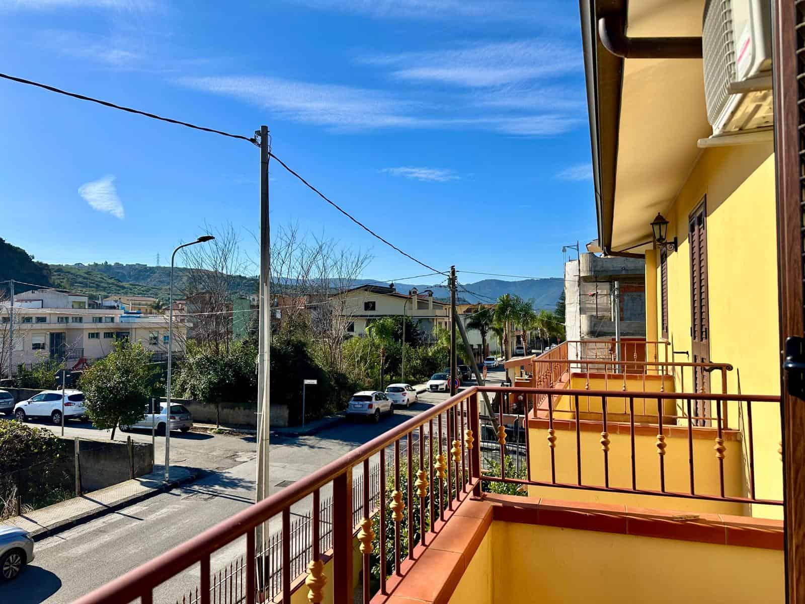 Vista balcone con strade, case, montagne e cielo azzurro, affaccio su strada di città soleggiata in zona residenziale con palme e edifici moderni, atmosfera tranquilla e luminosa in zona urbana.