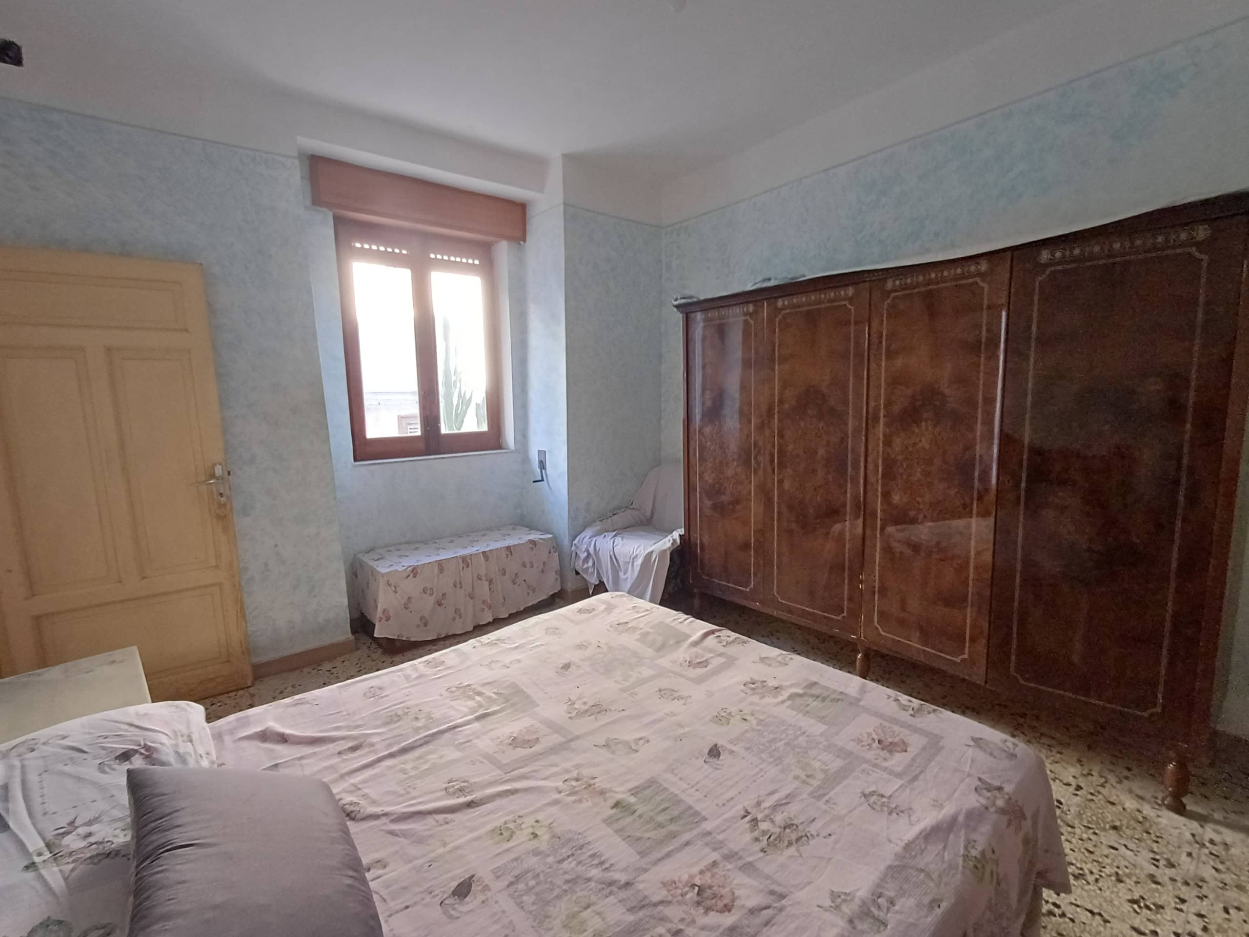 Ampia camera da letto con letto matrimoniale, armadio antico in legno, finestra con vista e arredamento classico in stile vintage, ideale per affitti turistici e soggiorni confortevoli.