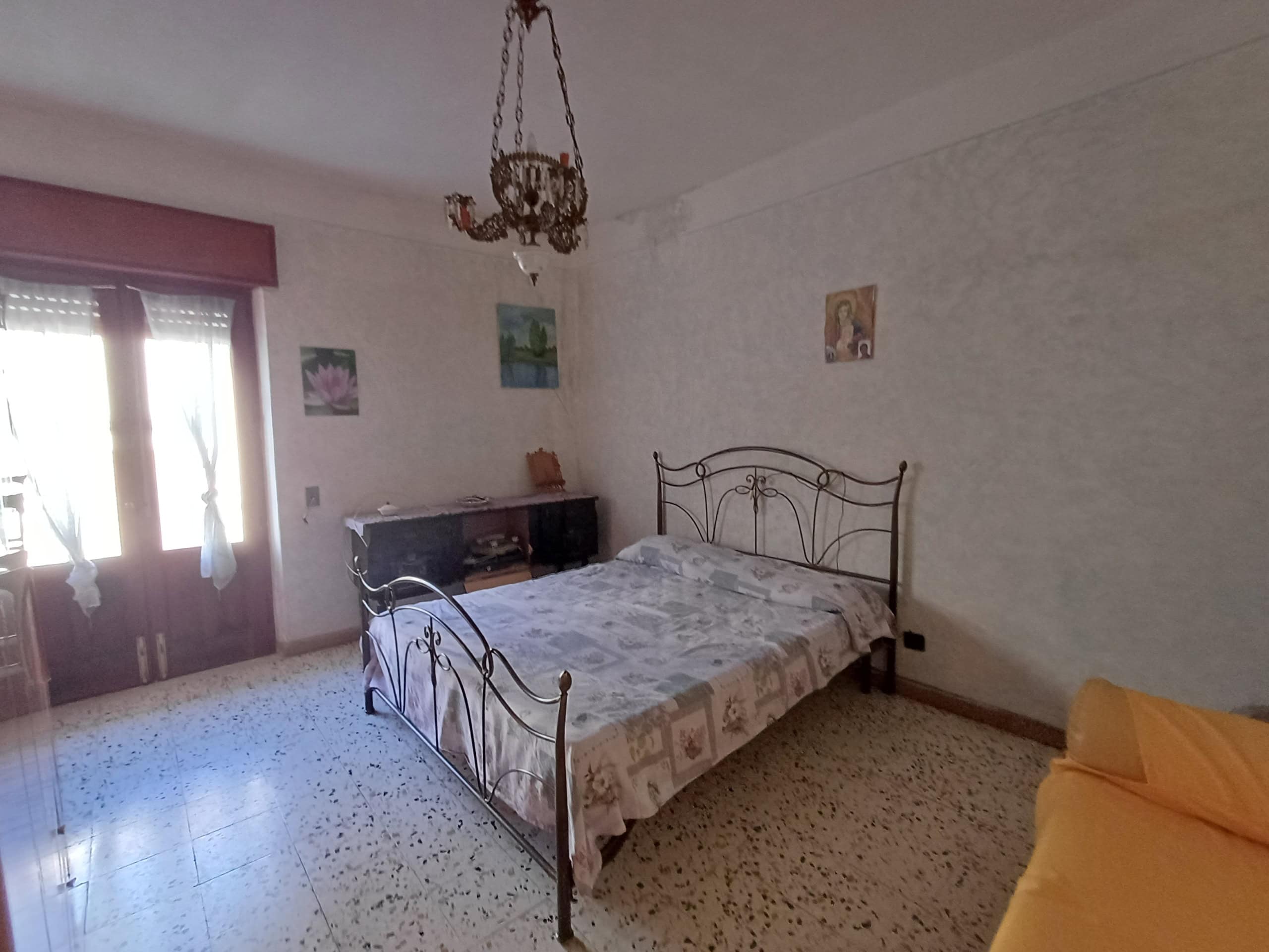 Letto matrimoniale con testiera in ferro battuto in camera da letto, arredamento classico, pareti colorate e lampadario antico, zona giorno luminosa con balcone, ideale per residenze tradizionali.