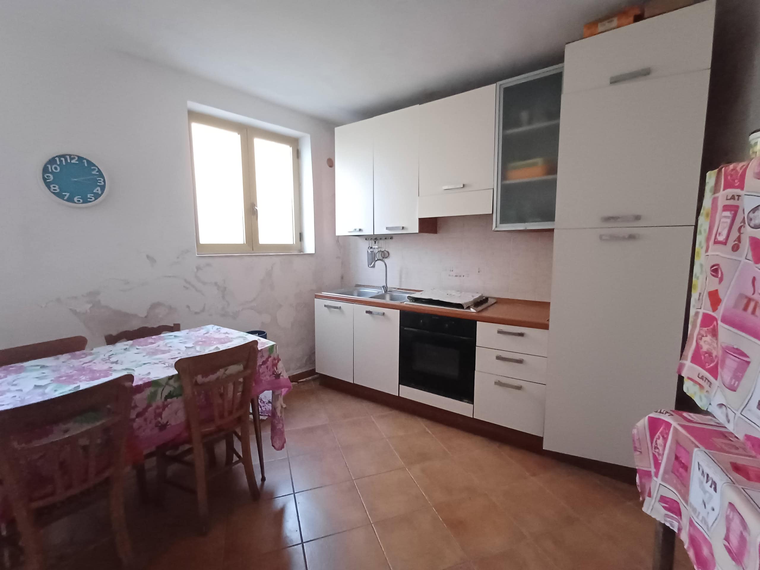 Lavanderia e cucina abitabile in appartamento con finestra, mobili bianchi e piano in legno, forno e frigorifero decorato con adesivi, tavolo con tende a fiori, stile semplice e funzionale.