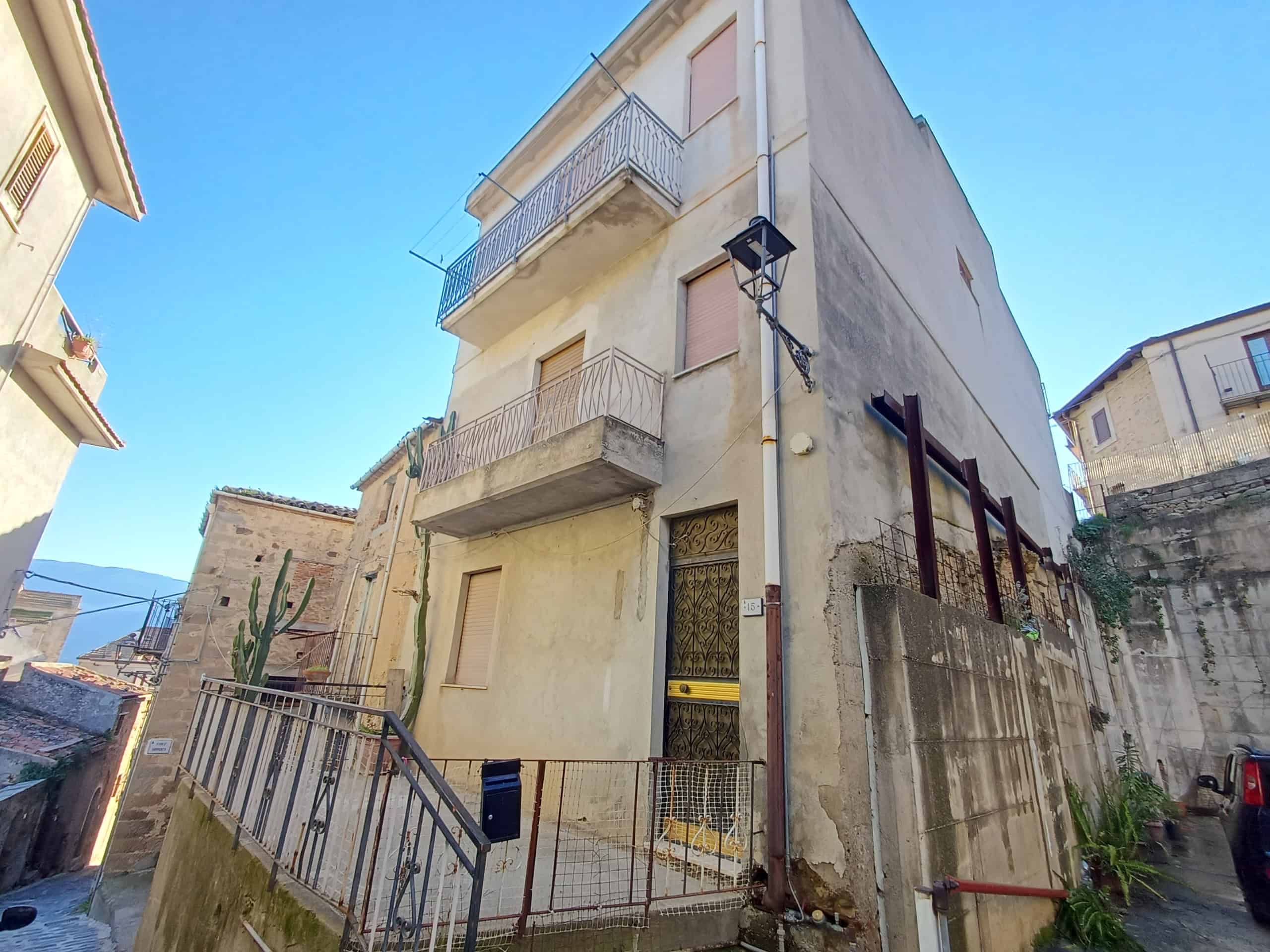 Vecchia casa multifamiliare in pietra e cemento con balconi, grate alle finestre e percorso in salita in un quartiere di montagna. Edificio storico con pareti esterne osservate sul lato strada e del vicolo.