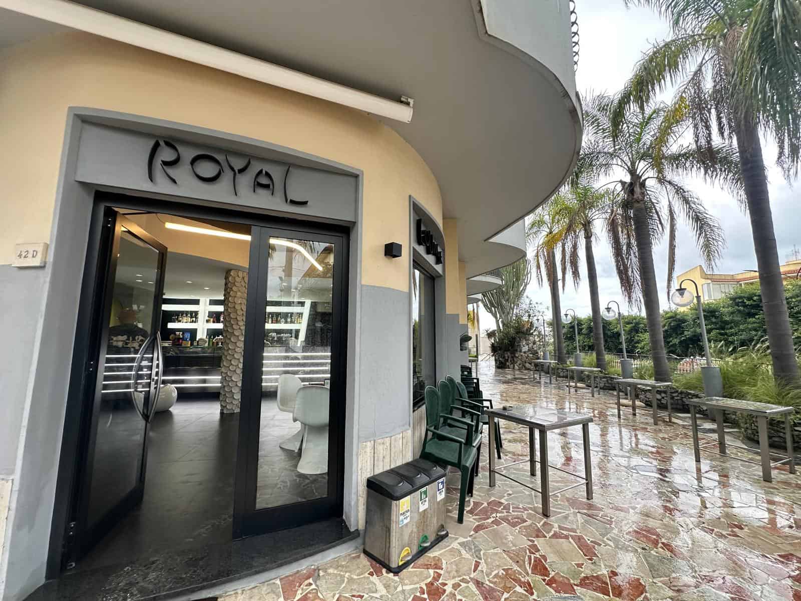 Nella foto si vede l’ingresso di un bar chiamato Royal, con un’ampia area esterna con sedie e tavoli, palme e un’atmosfera tropicale, ideale per relax e svago. Perfetto per chi cerca locali eleganti e atmosfere accoglienti in zone di città o resort.