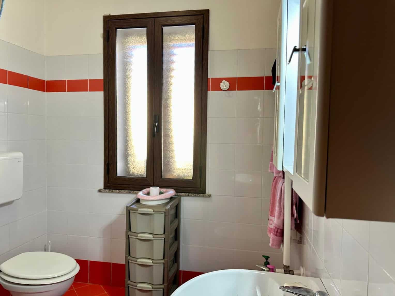 Bagno con finestra in legno e piastrelle bianche con dettaglio rosso, lavabo e mobiletto per riporre oggetti, ambiente funzionale e luminoso NextCasa.