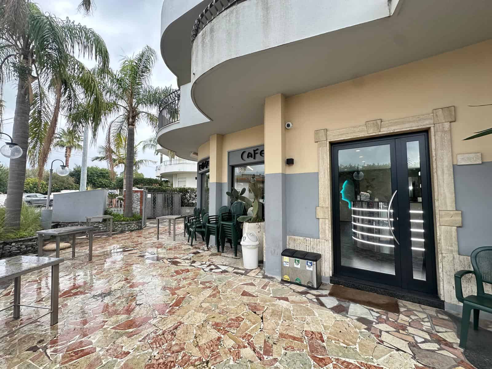Ingresso di un locale commerciale NextCasa con arredamento moderno e spazio esterno con panchine e palme, ideale per investimento immobiliare e attività commerciali a Milano.