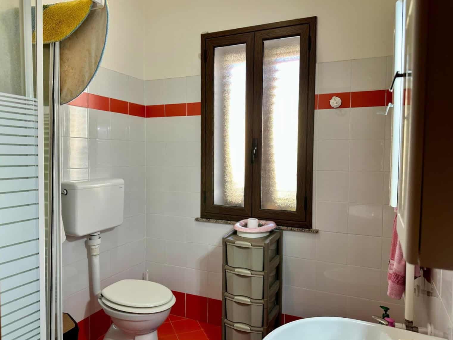 Bagno moderno con tenda doccia, finestra in legno, sanitari bianchi e pareti rivestite con piastrelle biene bianche e rosse, arredi pratici e spazio ottimizzato.
