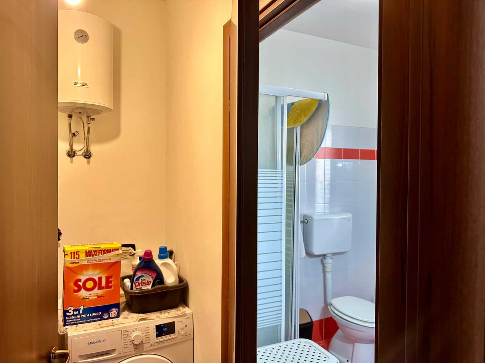 Lavatrice e scopa in lavanderia accanto al bagno con cabina doccia e WC, ambiente pulito e funzionale, ideale per vivere comodamente in casa NextCasa.
