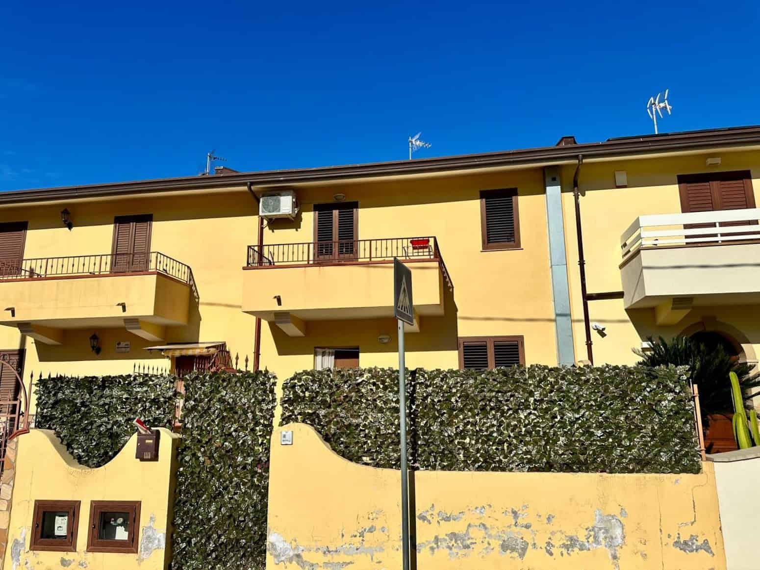 Ampio edificio residenziale color giallo con balconi, finestre con tapparelle e piante ornamentali, sotto un cielo azzurro, stile italiano, vicino a servizi e aree verdi, immobiliare NextCasa.