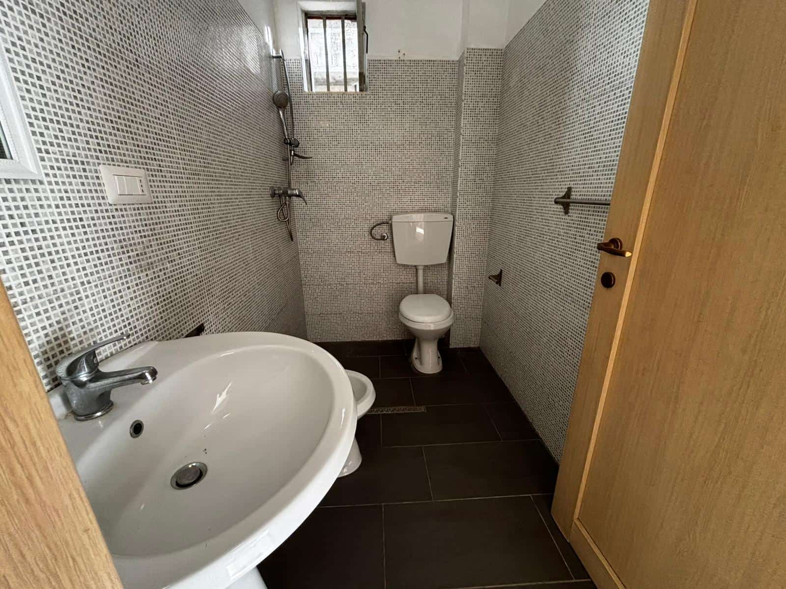 Bagno in appartamento con lavabo, WC e doccia, pavimento in piastrelle scure, pareti rivestite con mosaico, luce naturale, stile funzionale e moderno.