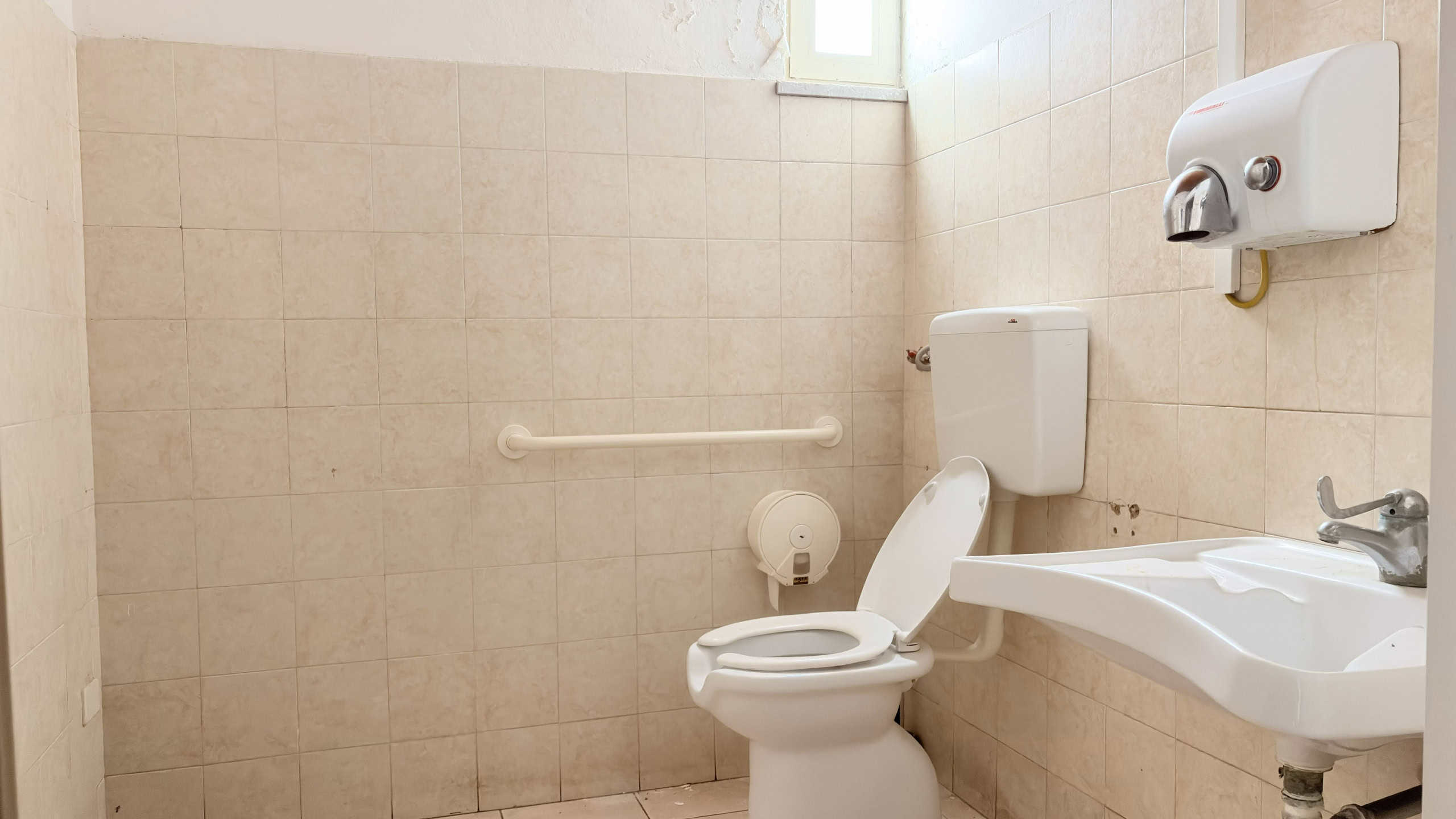 Pulizia bagno accessibile con sedia a rotelle, WC e lavabo in ambiente neutro, illuminato naturalmente, per servizi di assistenza domiciliare e comfort.