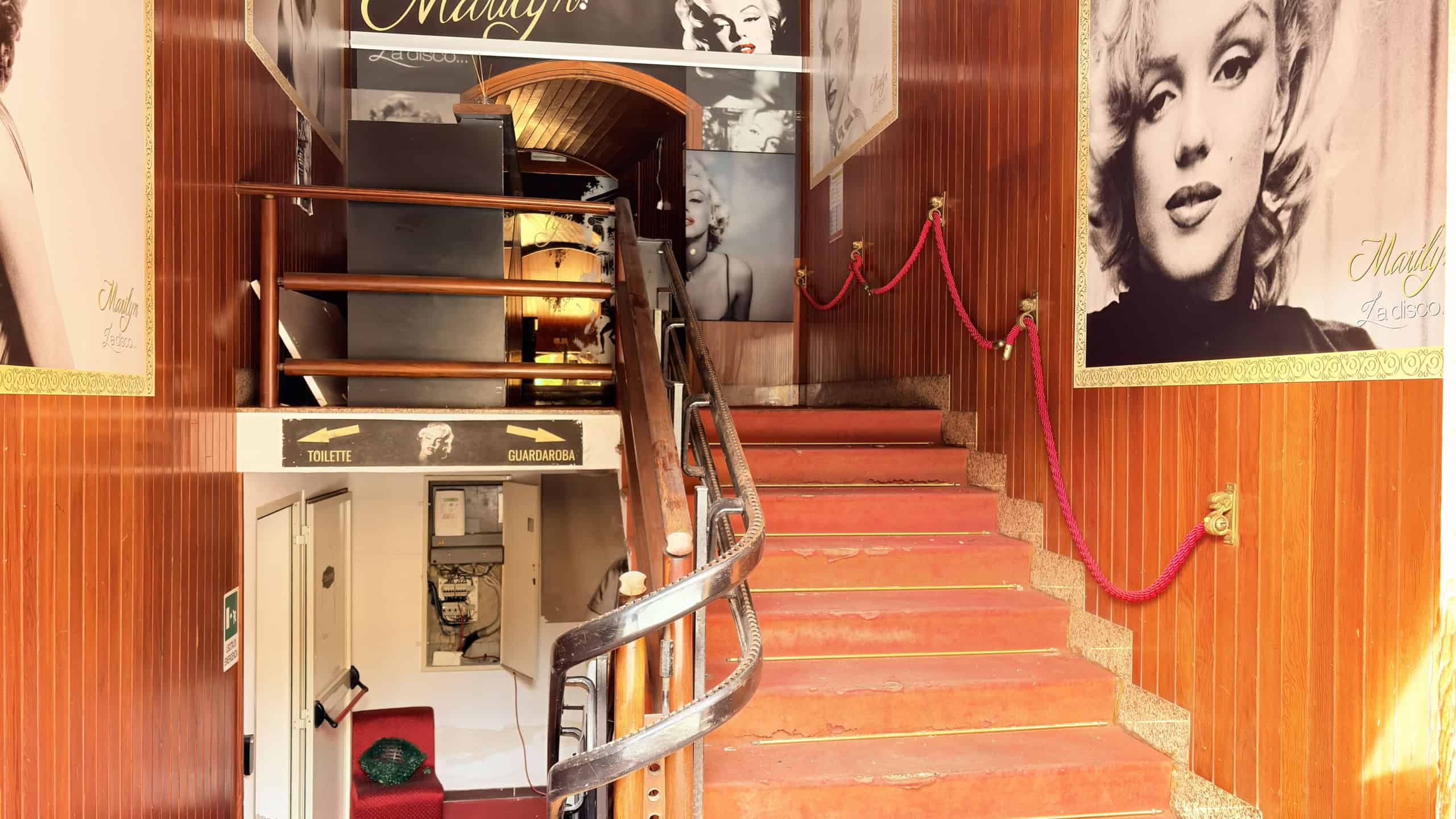 Elegante ingresso di un locale in stile vintage con pareti rivestite in legno, poster iconici di Marilyn Monroe e una scaletta in legno e metallo, atmosfera rétro e decorazioni di classe.