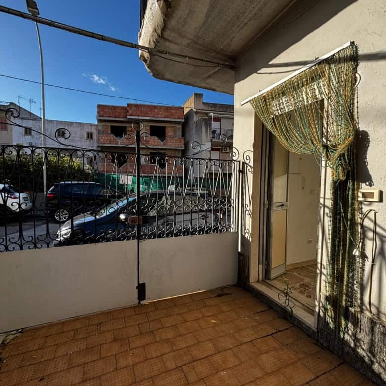 Balcone di casa in zona urbana con vista su parcheggio e edifici circostanti, recinzione in ferro battuto, portafinestra con tenda a filamenti decorativa, luce naturale e atmosfera accogliente.