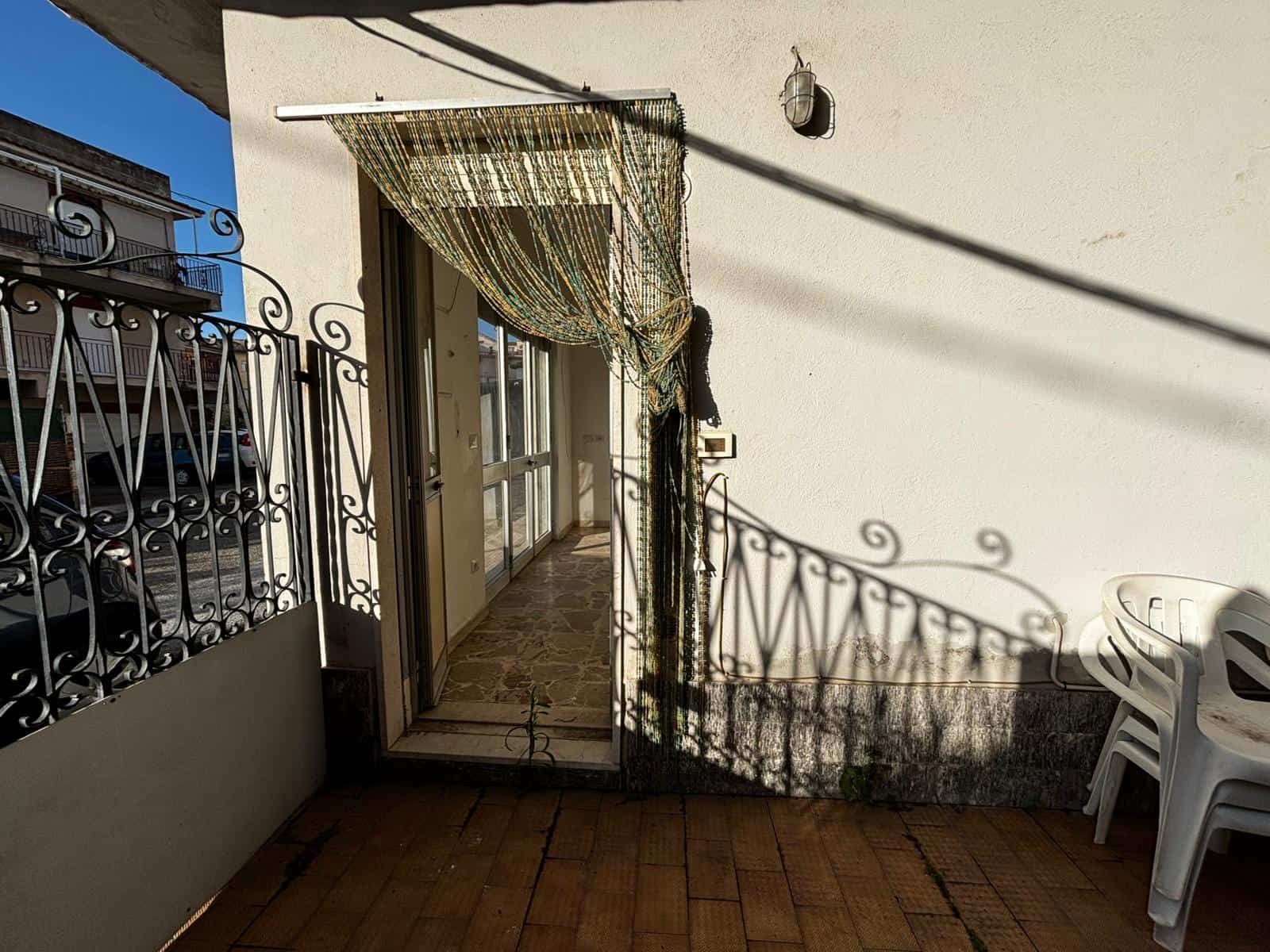 Balcone con ringhiera in ferro battuto, porta finestra con tendaggio di stoffa, arredamento semplice con sedie di plastica bianche, pavimento in piastrelle in ceramica, in una casa vicino al centro urbano.