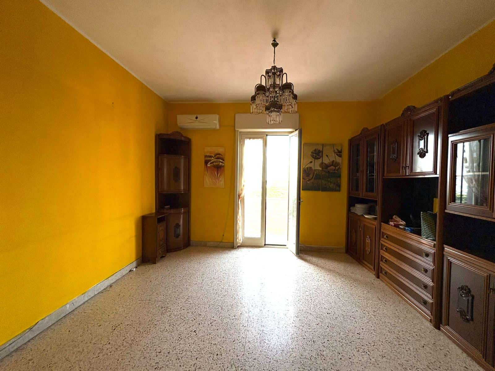 Antico soggiorno con pareti gialle, mobili in legno scuro, lampadario a cristalli, grande porta-finestra che dà accesso al balcone, ideale per rinnovare un'abitazione con stile classico.