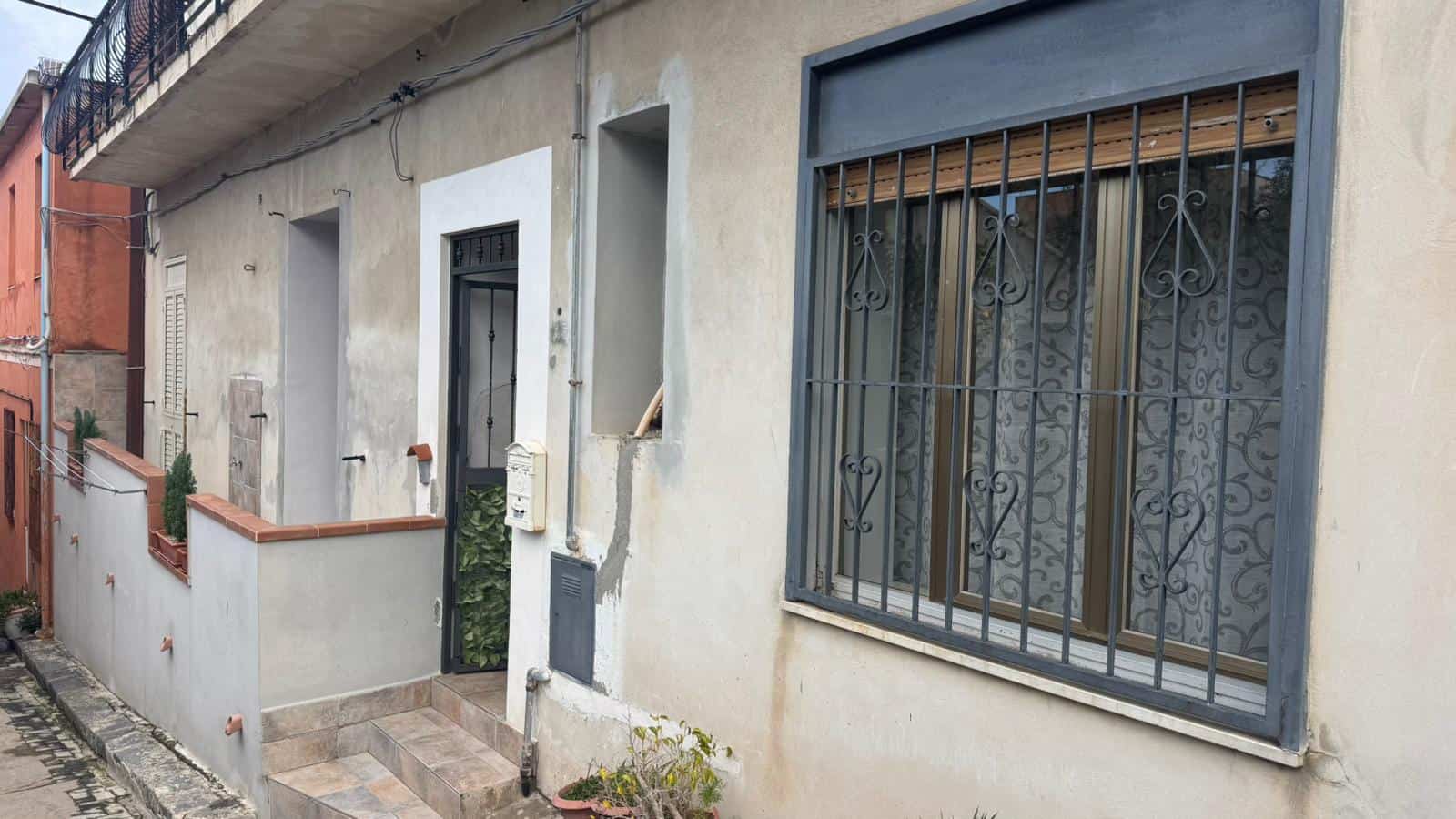 1. Nascosta casa con pareti esterne in stucco grigio, finestre con grate decorative e piccola area esterna con piante e gradini in ceramica.