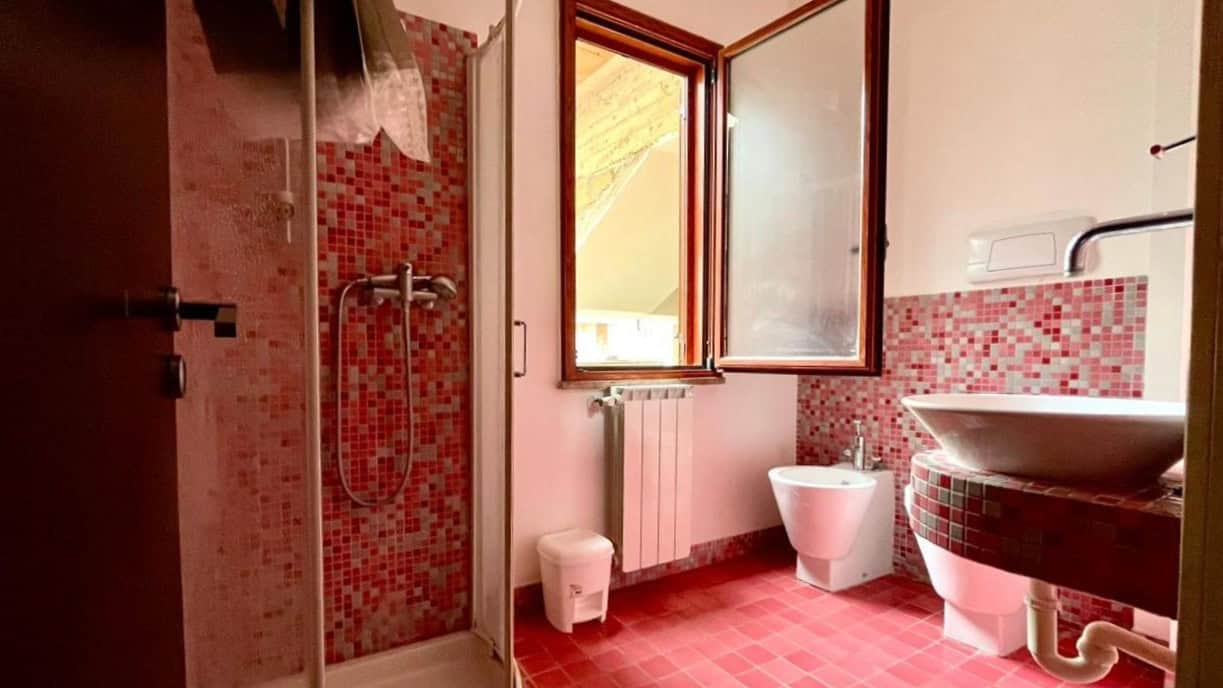 Doccia con piastrelle rosse, finestra aperta, lavabo moderno e bidet in un bagno luminoso e accogliente, ideale per case vacanze e proprietà di lusso, perfetta per chi cerca comfort e stile.