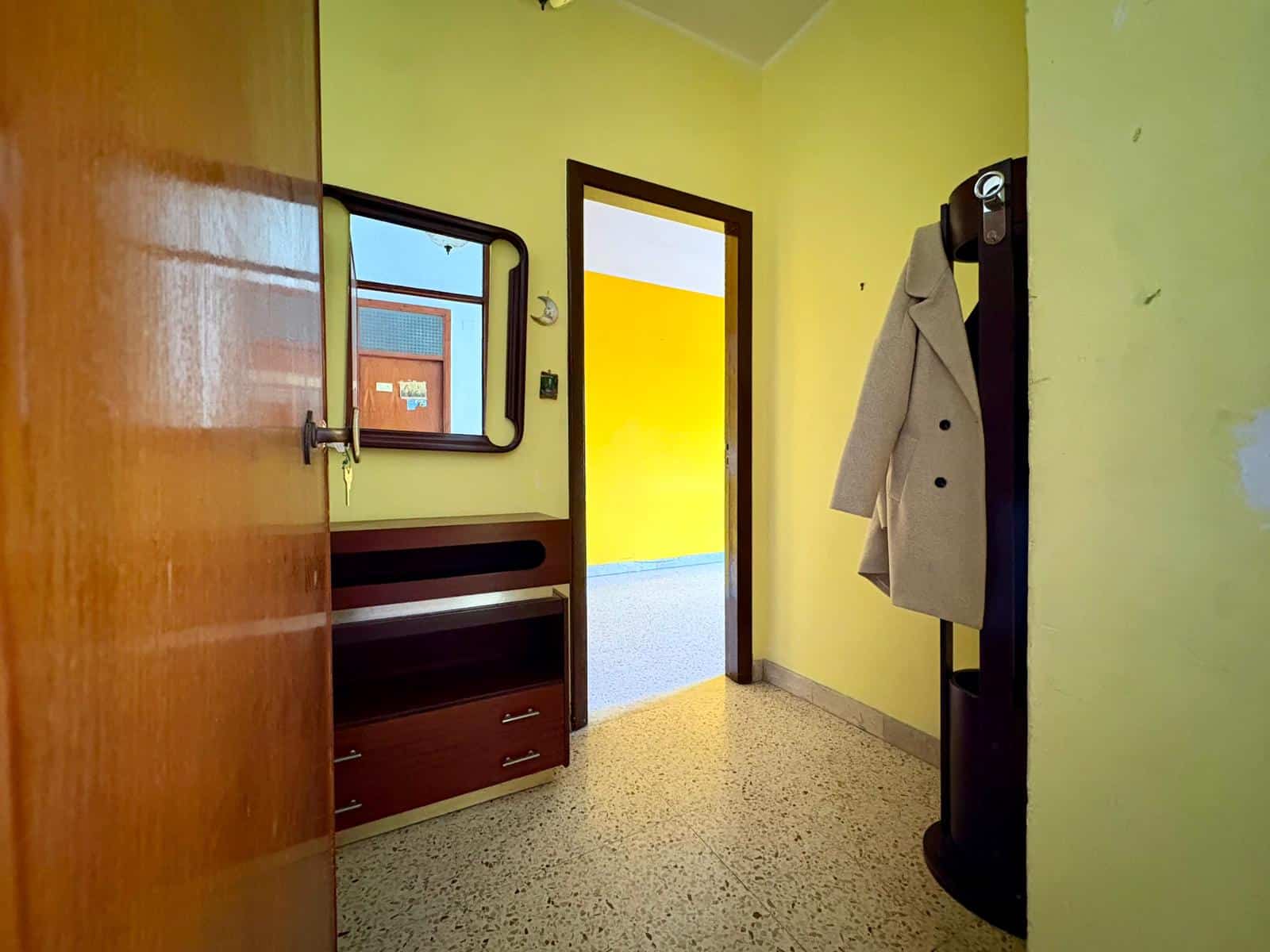 Ingresso di appartamento con pareti gialle, specchio e mobile contenitore, porta d'ingresso blu sullo sfondo, sistema di accoglienza con cappotto beige appeso, pavimento in terrazzo.