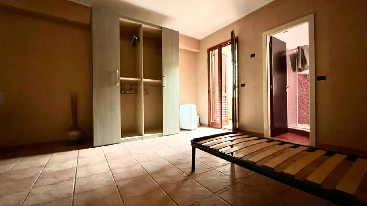 Armadio a due ante in camera da letto con spazio per vestiti e accessori, vicino a porta con finestra che dà sul balcone. Camera luminosa e spaziosa con pavimento in piastrelle e arredamento minimale.
