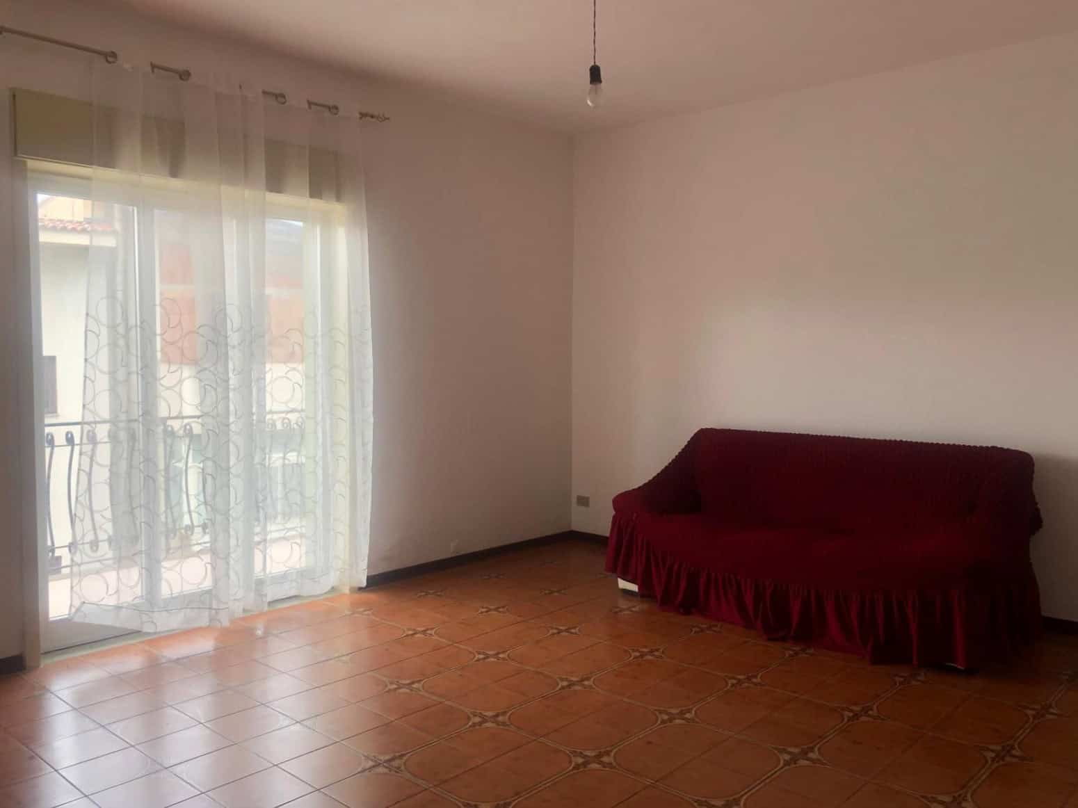 Soggiorno luminoso con accesso a balcone, pareti bianche, pavimento in ceramica decorata, divano rosso coperto, tende leggere, atmosfera accogliente e spaziosa.