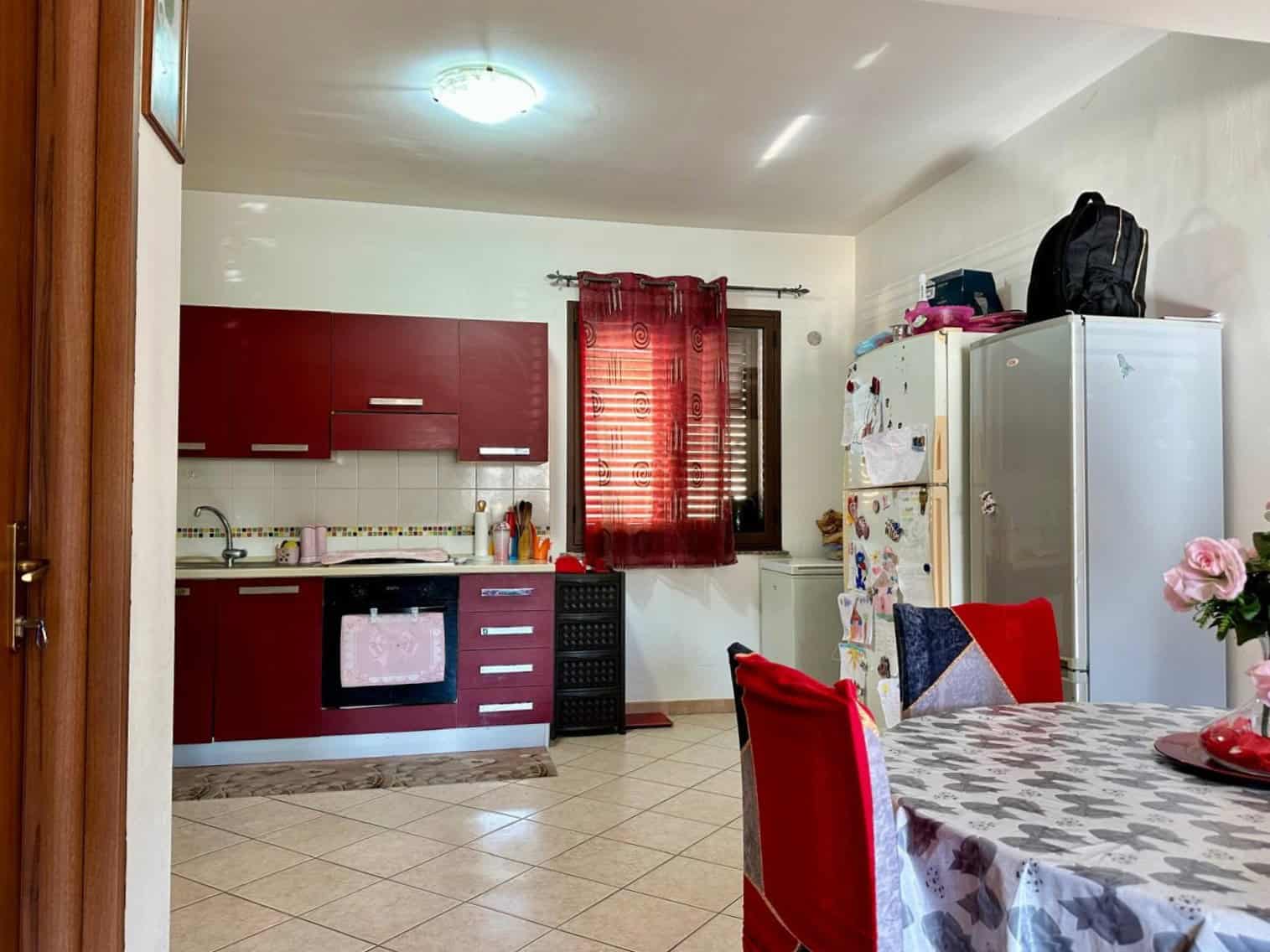 Cucina abitabile con mobili rossi e porta finestra con tenda rossa, zona pranzo con tavola decorata, frigorifero bianco, detersivi e accessori da cucina, ambiente luminoso e accogliente.