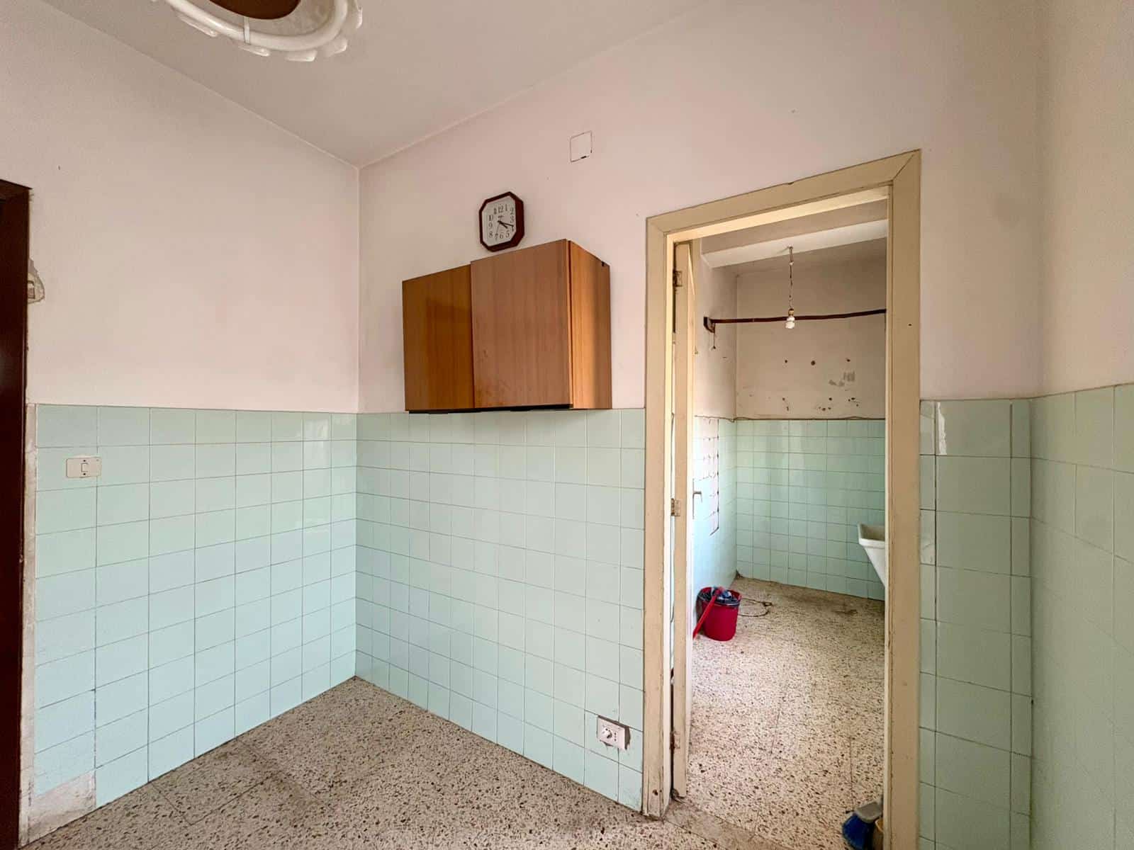 Stanza da cucina abitabile con rivestimento in piastrelle verde acqua, armadietto in legno e porta che conduce a un locale adiacente con lavandino e spazio per lavatrice, arredo semplice e funzionale.