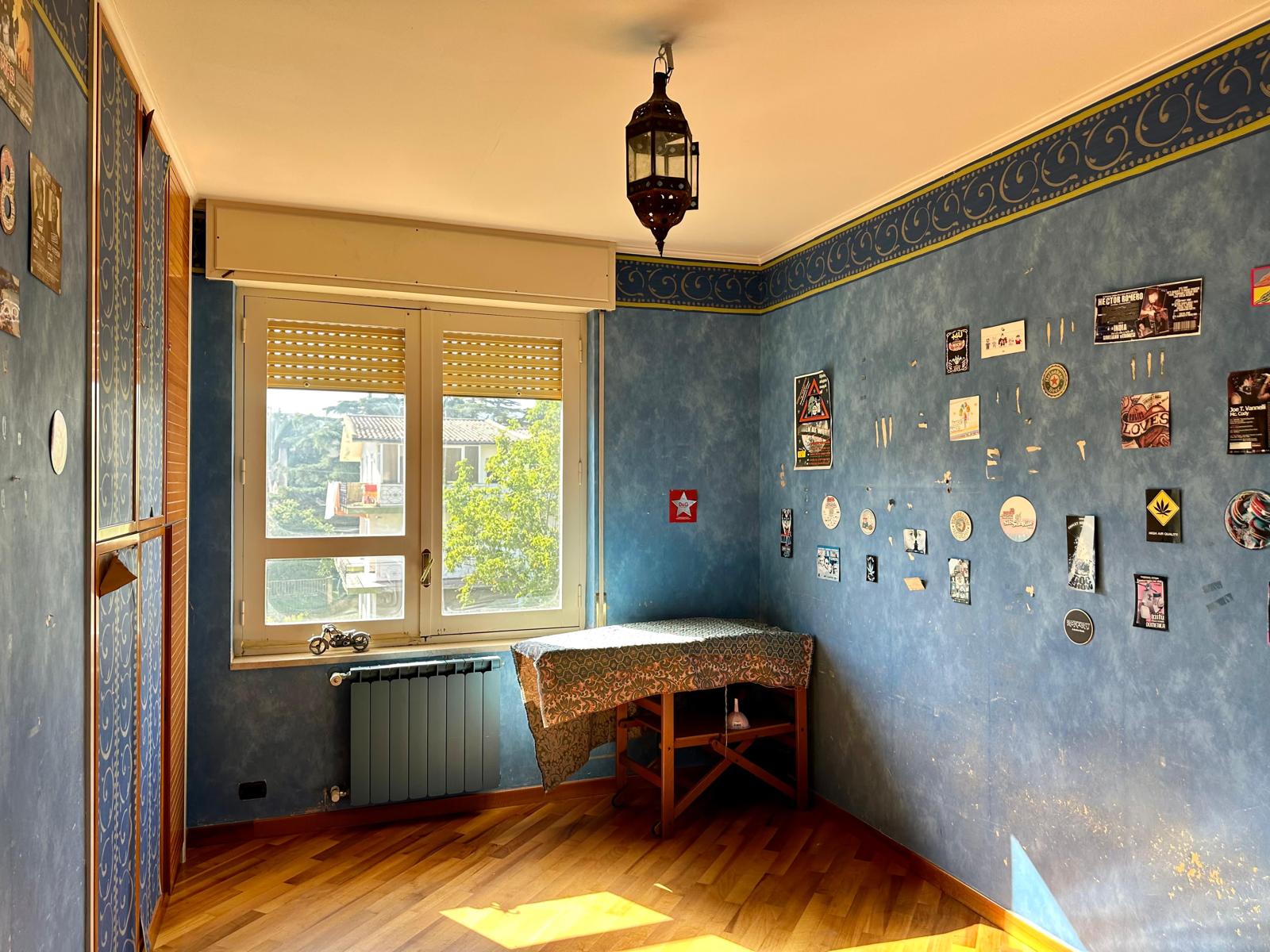 Vecchio soggiorno con pareti blu, decorazioni e adesivi, grande finestra con vista su giardino, riscaldamento a radiatore, arredo semplice con tavolo e lampadario rustico in stile marocchino.